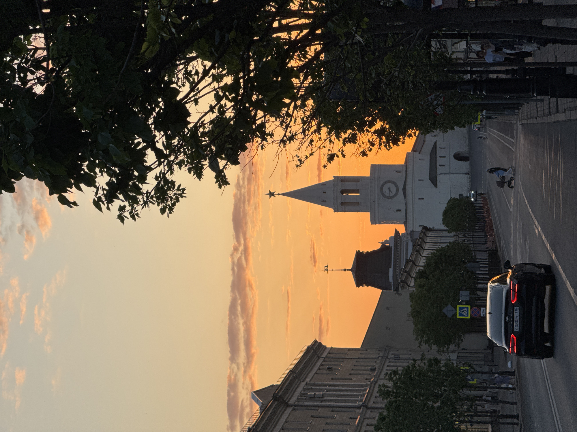The image depicts a street in Kazan, Russia, at sunset. In the background is the Soyembika Tower of the Kazan Kremlin, recognizable by its distinctive leaning silhouette and the star atop its spire. The sky is a warm, orange hue due to the setting sun. A black car is in the foreground, driving along the street. On the sidewalk, there is a person walking with a stroller. A couple is standing at the side. The street is lined with buildings, and trees are visible on both sides. A pedestrian crossing sign is also visible next to the car.