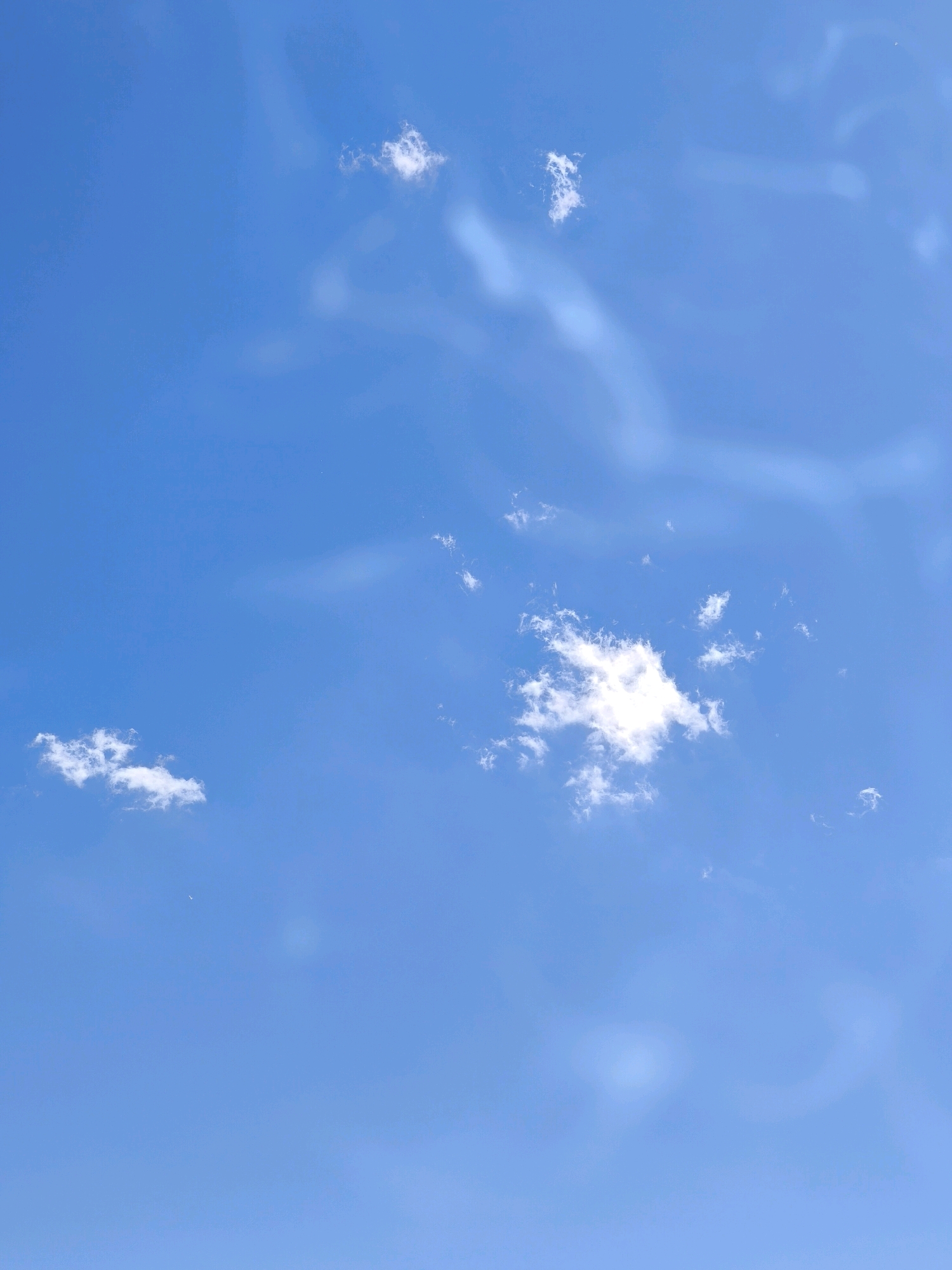 The image is a vertical, eye-level shot of a bright blue sky with scattered white clouds. There are no people, buildings, or any other discernible man-made objects visible. The scene is natural and serene, with the wispy clouds appearing as delicate brushstrokes against the vast expanse of blue. The lighting suggests a clear day, likely during daylight hours. The environment is solely the sky. There is no visible indication of the city of Karachayevsk, Russia, or any specific location on Earth.