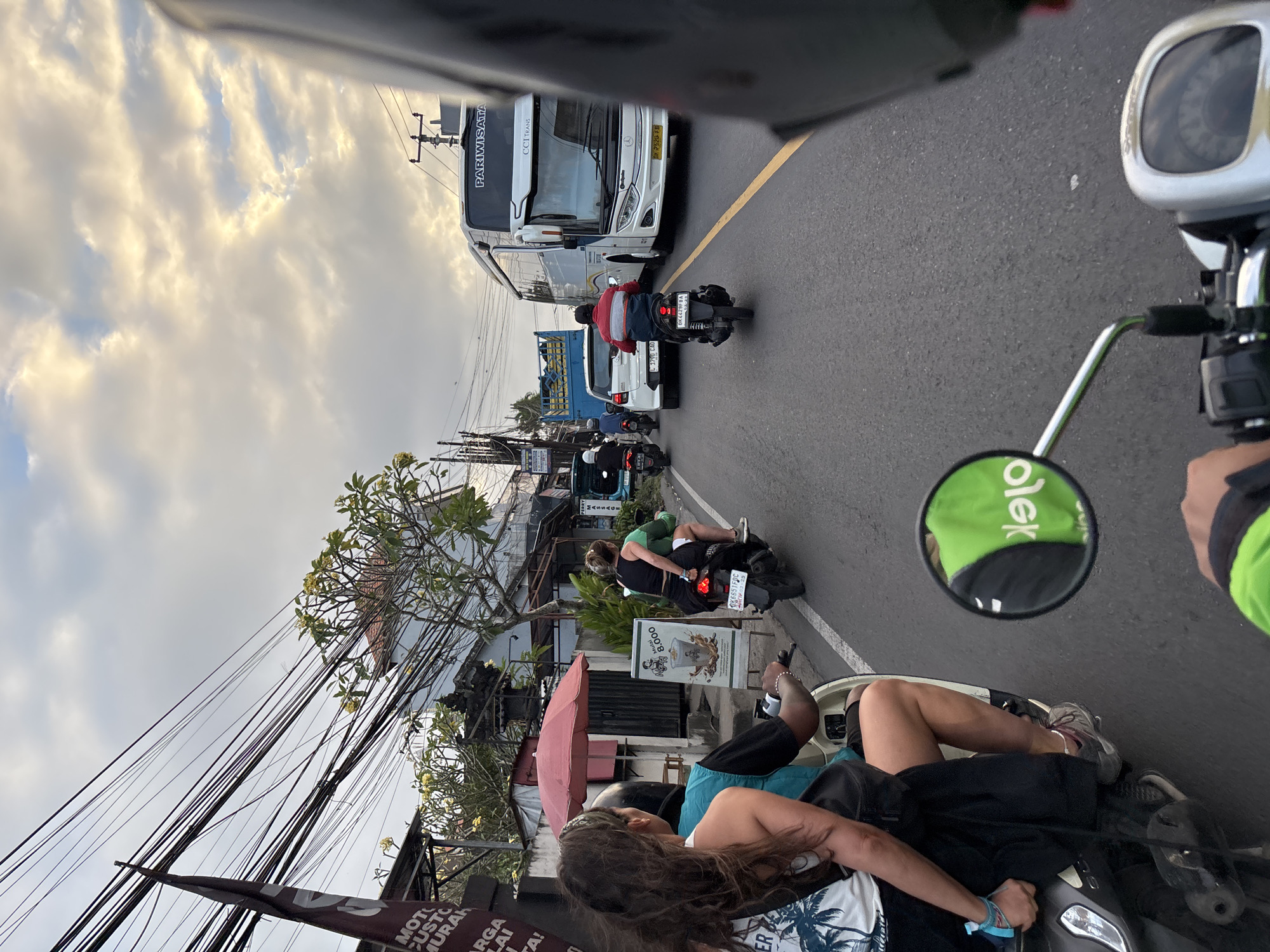 A POV shot from a motorcycle in Kuta Selatan, Indonesia shows a busy street scene during daylight. A woman with long brown hair and a blue tank top is a passenger on the back of the motorcycle. She is wearing black shorts and a white t-shirt with a palm tree design. The driver of the motorcycle is wearing a blue tank top and a black helmet. In front of them, a woman in a black tank top and green vest is riding a black motorcycle. Further ahead, a white passenger bus with "PARIWISATA" and "CCI TRANS" written on it is driving on the road. A white car and a blue truck are also visible on the road. Power lines and trees with yellow flowers are seen on the side of the road. A sign with "Mulal Rp 8.000" is also displayed on the side of the road. The sky is cloudy with patches of sunlight.