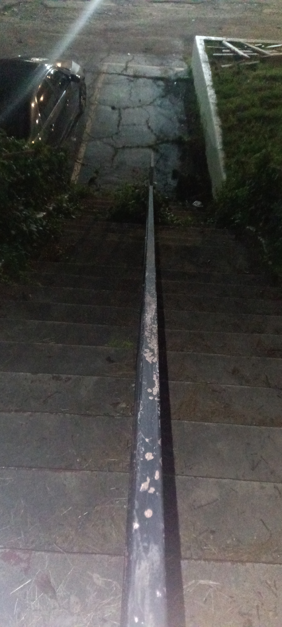 The image depicts an outdoor staircase leading downwards. The steps appear to be made of stone or concrete. A metal handrail runs down the center of the staircase. Vegetation is visible on either side of the staircase. At the top of the stairs, a parked car is partially visible to the left, and a patch of grass enclosed by a wall is on the right. The lighting suggests it is nighttime or a dimly lit area. This location is in the city of Fort Smith, United States of America.