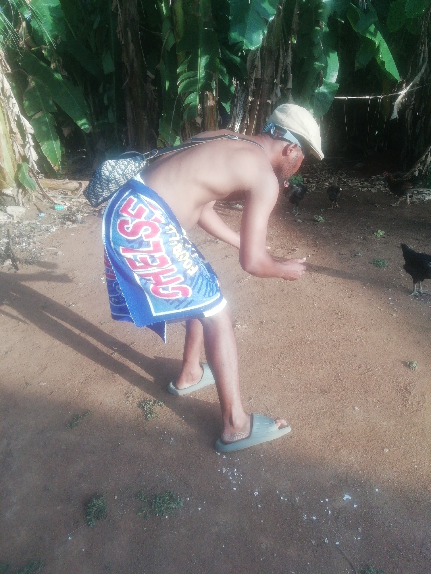 A shirtless man wearing Chelsea FC shorts, a cap, and slides is crouching in a dirt yard, appearing to scatter something for several chickens. The background features lush, tropical foliage. The location appears to be rural.