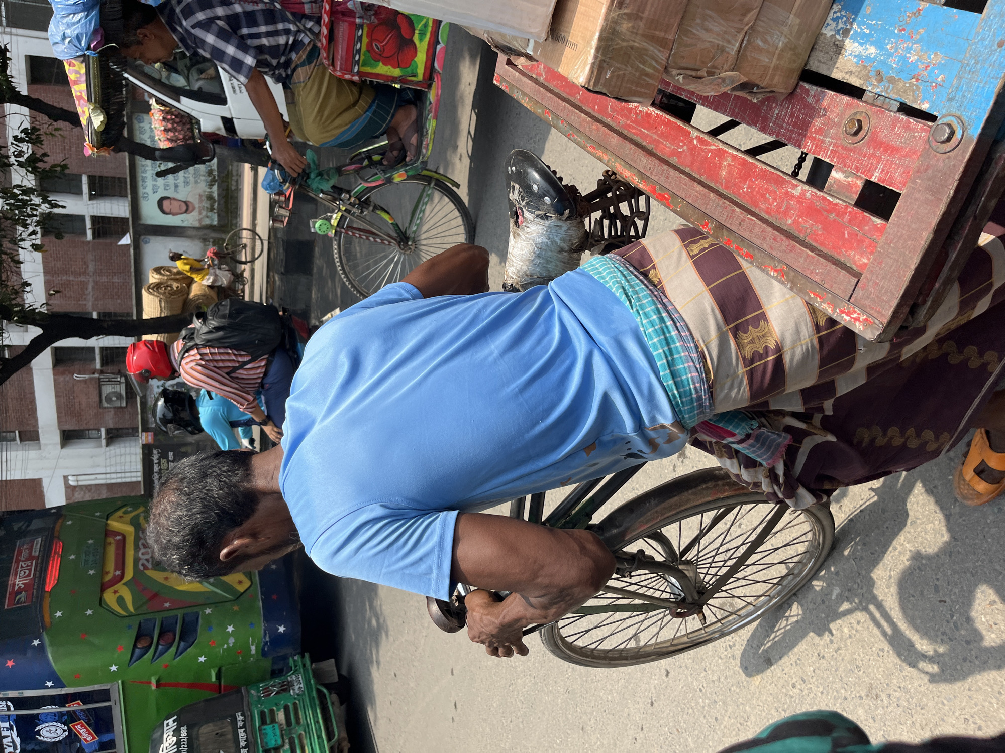 The image depicts a street scene in Dhaka, Bangladesh. A man in a blue shirt and lungi (traditional garment) is pedaling a bicycle laden with a red and blue wooden cargo carrier. The bicycle is positioned beside a green and multi-colored bus. In the background, another man is seen riding a similar bicycle laden with items. The setting is an urban street with buildings and other vehicles visible in the distance.