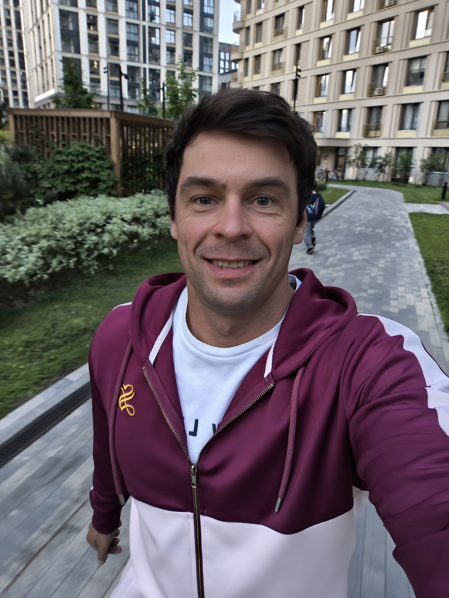 A man is taking a selfie outdoors. He is wearing a maroon and white zip-up hoodie and a white t-shirt. Behind him is a paved walkway with grass and shrubbery on either side. In the background are modern apartment buildings. The scene appears to be in Moscow, Russia, as the architectural style of the buildings is characteristic of contemporary Moscow development. Another person is walking behind him in the background.