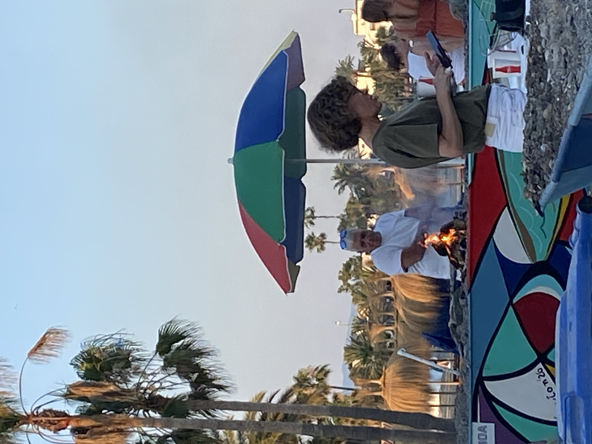 The image depicts an outdoor scene, possibly a beach or boardwalk, with several people present. In the foreground, a colorful, multi-panel umbrella is prominently featured. A woman with curly hair is visible in profile, wearing a green shirt and white shorts, and holding a tablet or phone. Further back, a man wearing a white shirt and sunglasses is standing near a small fire. The background shows palm trees and buildings, suggesting a warm, sunny climate. The lighting indicates it could be late afternoon or early evening. The scene appears relaxed and casual, possibly a social gathering or a vendor setting up for an event. A vibrant, abstract mural is visible on a wall to the right, with some text inscribed on it, though it is partially obscured. The overall atmosphere is lively and summery.