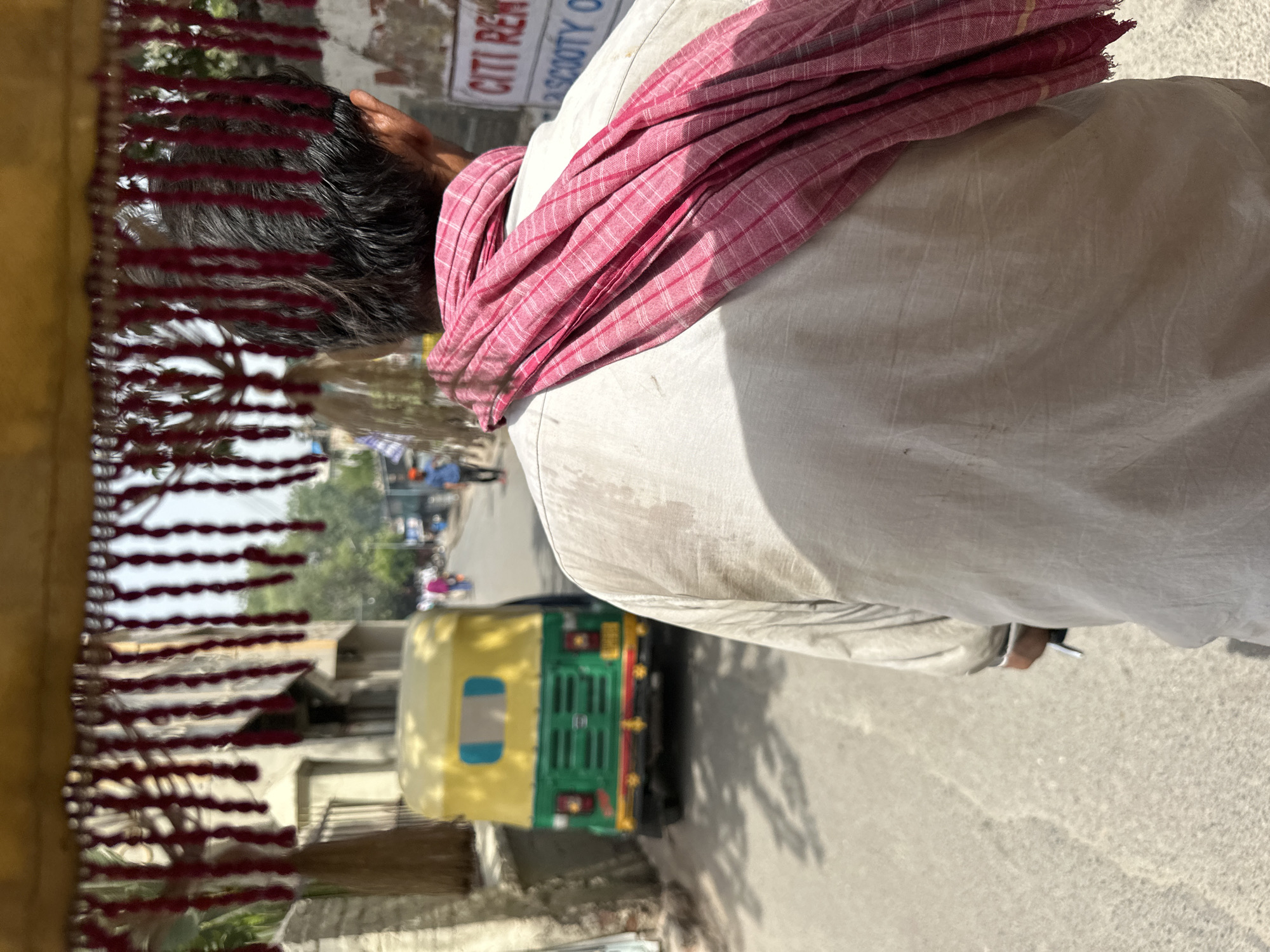 The image shows a view from inside a motorized rickshaw in Varanasi, India. The driver, seen from the back, is wearing a white shirt and a pink-and-white checkered cloth draped over his shoulder. The rickshaw's decorative fringe is visible in the foreground. An auto-rickshaw is next to the subject rickshaw. A building with signage is visible in the background. The view suggests movement along a street.