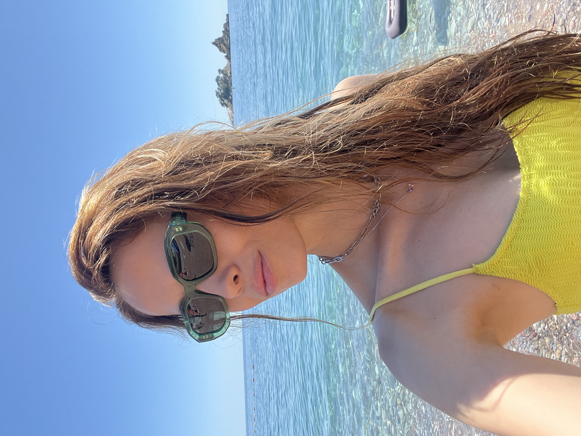 A bright, sunlit, tilted selfie captures a young woman enjoying a sunny day by the sea. The woman is the primary subject, positioned on the left side of the frame, looking towards the viewer with a subtle, relaxed smile. She has long, light brown hair, which appears slightly wet, flowing over her shoulder. She wears fashionable, rectangular green-framed sunglasses, reflecting the clear blue sky and her surroundings. A delicate silver chain necklace is visible around her neck.
She is dressed in a vibrant, textured yellow bikini top. The background is dominated by incredibly clear, turquoise-blue water, through which light-colored pebbles on the seabed are distinctly visible, indicating shallow and pristine conditions. A rocky island or headland is discernable on the distant horizon, set against a vast, cloudless blue sky. The scene conveys a relaxed, summery atmosphere at a picturesque coastal location, likely Petrovac centar, Montenegro. The time of day is clearly daytime, given the bright sunlight. There is no visible text in the image.