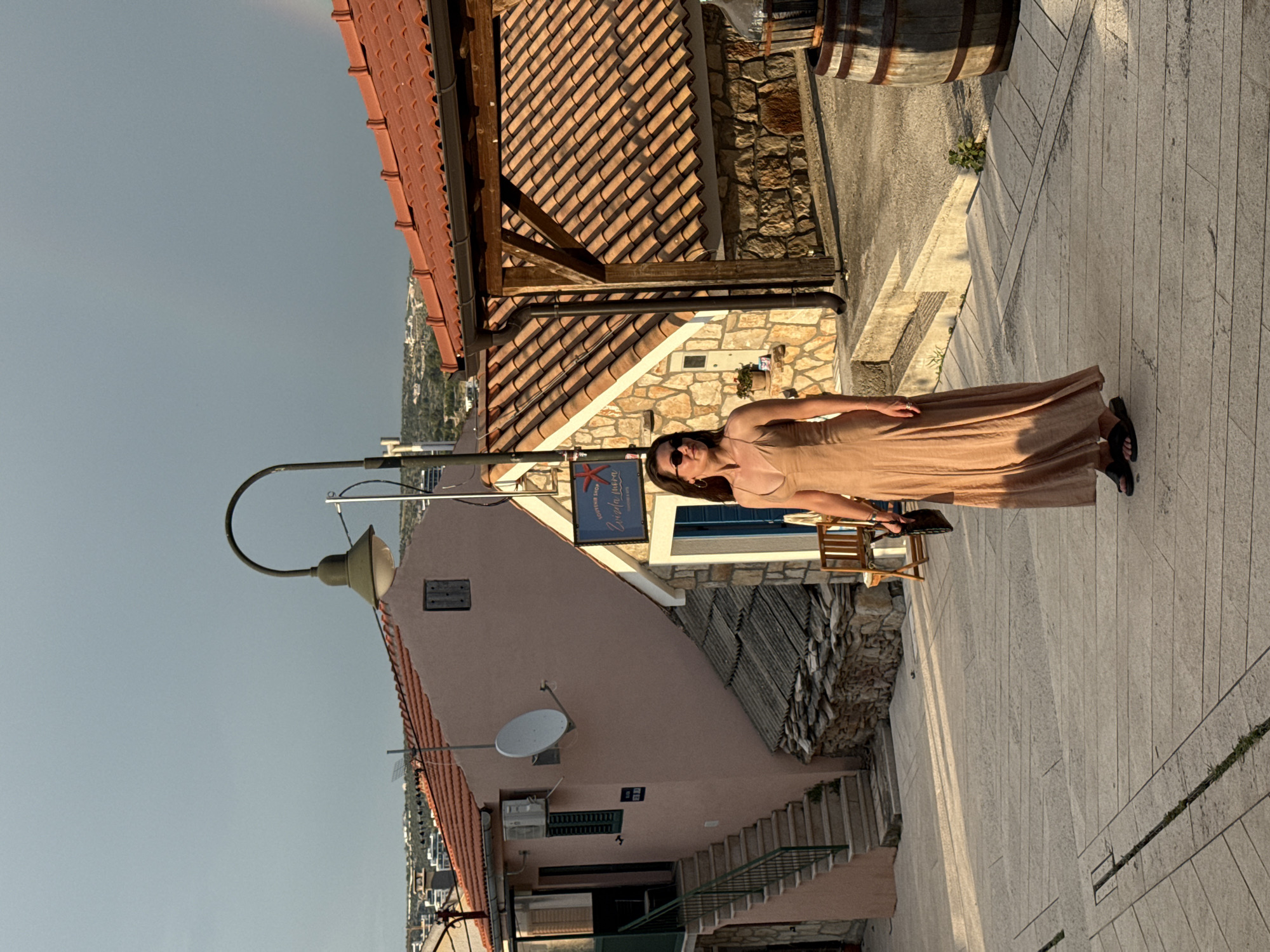 A woman wearing a tan, sleeveless maxi dress and black sandals stands on a paved street in Primošten, Croatia. She is looking to her left and holding a dark clutch bag. The street is paved with large, rectangular stones. To her left is a pale pink building with a satellite dish and air conditioning unit mounted on the exterior. A set of outdoor stairs with a green railing leads up to the building. To her right, there is a stone building with a wooden overhang and a red-tiled roof. A sign above the entrance reads "SOUVENIR SHOP Zvezda mora". A wooden barrel and a large glass bottle are visible next to the stone building. The background shows more buildings and distant hills under a cloudy, overcast sky. The sunlight casts long shadows on the street.