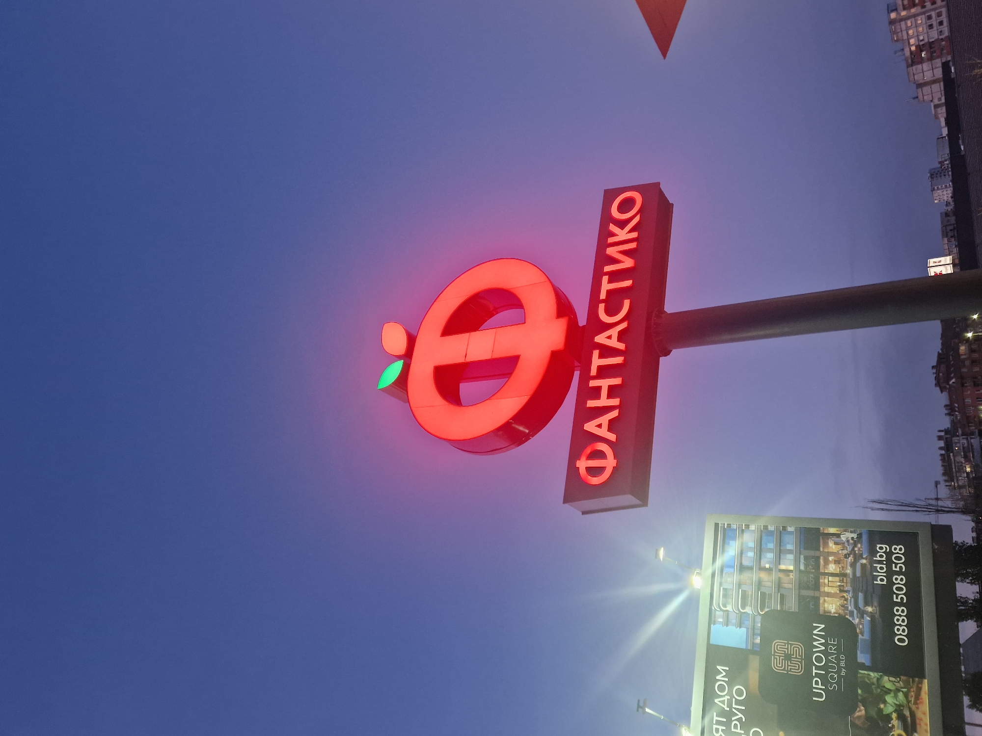 The image shows a large, illuminated red sign against a dark blue evening sky. The sign features a stylized red "e" logo with a small green leaf at its top left, alongside the Cyrillic word "ФАНТАСТИКО". This is the branding for the Bulgarian supermarket chain Fantastico. The sign is mounted on a dark pole.
Below the main sign, a brightly lit billboard is visible, featuring an advertisement for a real estate development. The billboard displays the text "UPTOWN SQUARE by BLD" and includes a website address "bld.bg" and a phone number "0888 508 508". Partial text, "ят дом" and "РУГО", is visible on the left side of the billboard, likely part of a Bulgarian phrase. The billboard also shows an image of a modern building.
In the background, to the right, faint outlines of buildings and structures can be seen against the fading light of dusk or early night. The sky transitions from a deep blue at the top to a slightly lighter hue towards the horizon, with a hint of purple-pink on the right. The overall scene suggests an urban or suburban setting in Bulgaria, given the Cyrillic text and the domain ".bg".