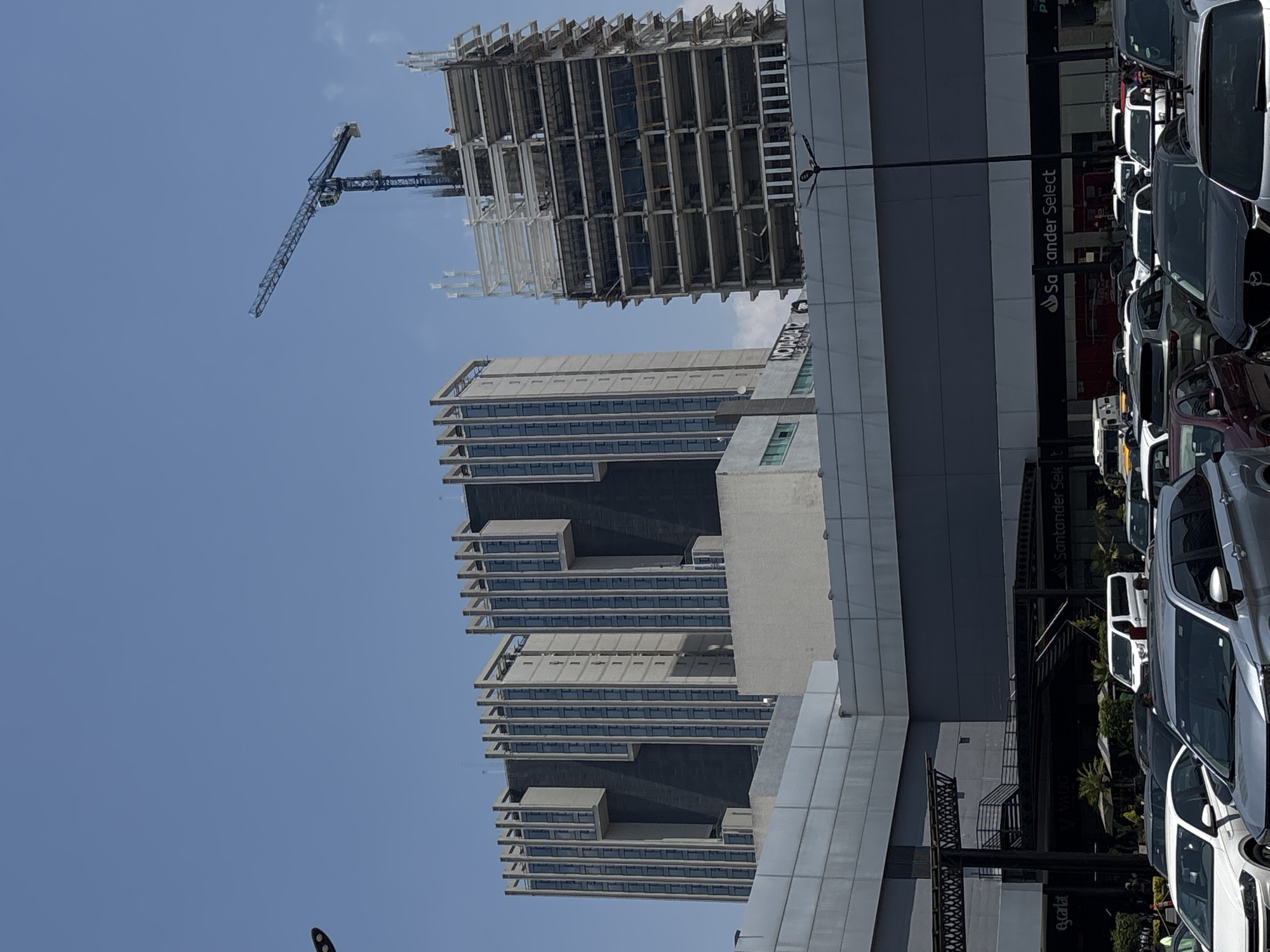 The image captures a construction site in San Bernardino Tlaxcalancingo, Mexico. A tall, unfinished building is being constructed with the help of a blue crane. A second, shorter building with a unique architectural design stands nearby. In the foreground, a street with parked cars is visible, and a Santander Select sign suggests a bank branch nearby. The sky is clear and blue.