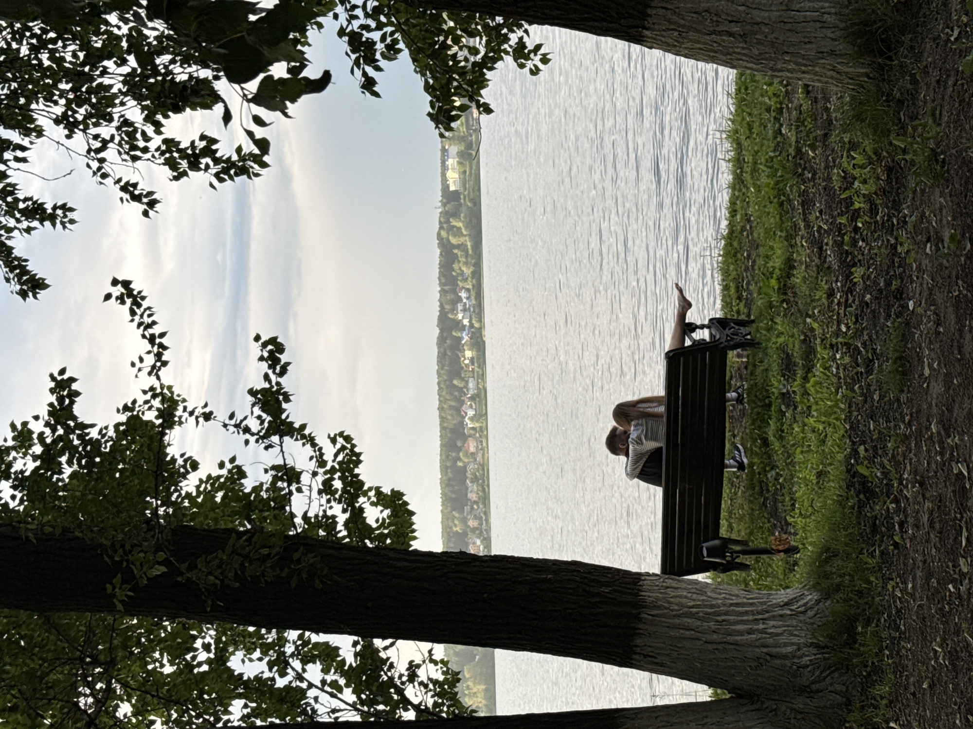 A couple is seated on a park bench facing a body of water, with a tree trunk in the foreground to the left and right, partially obscuring the view. The couple is embracing, with one person's arm around the other. The body of water is calm, with ripples on the surface, and a distant shoreline with buildings and trees can be seen across the water. The sky is overcast with some clouds. The ground in the foreground is covered with grass and fallen leaves. The scene appears to be in a park setting. The image location is Votkinsk, Russia, likely near the Votkinsk Reservoir, a popular recreational area in the city.