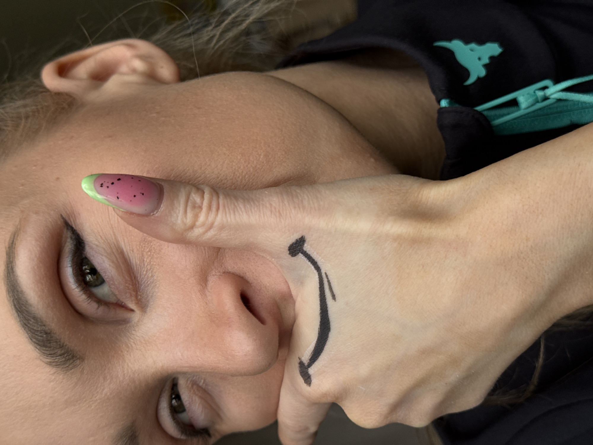 The image shows a person's face and hand. The person is fair-skinned with dark hair and eye makeup. The right hand is raised, covering part of the face, with a downward-facing smiley face drawn on the back of the hand. The thumb has a watermelon-themed nail design. The person is wearing a dark-colored top with a blue-green zipper. The location is not discernible from the image. The image does not provide enough context to determine if it was taken in the city of Chaikovsky, Russia.
