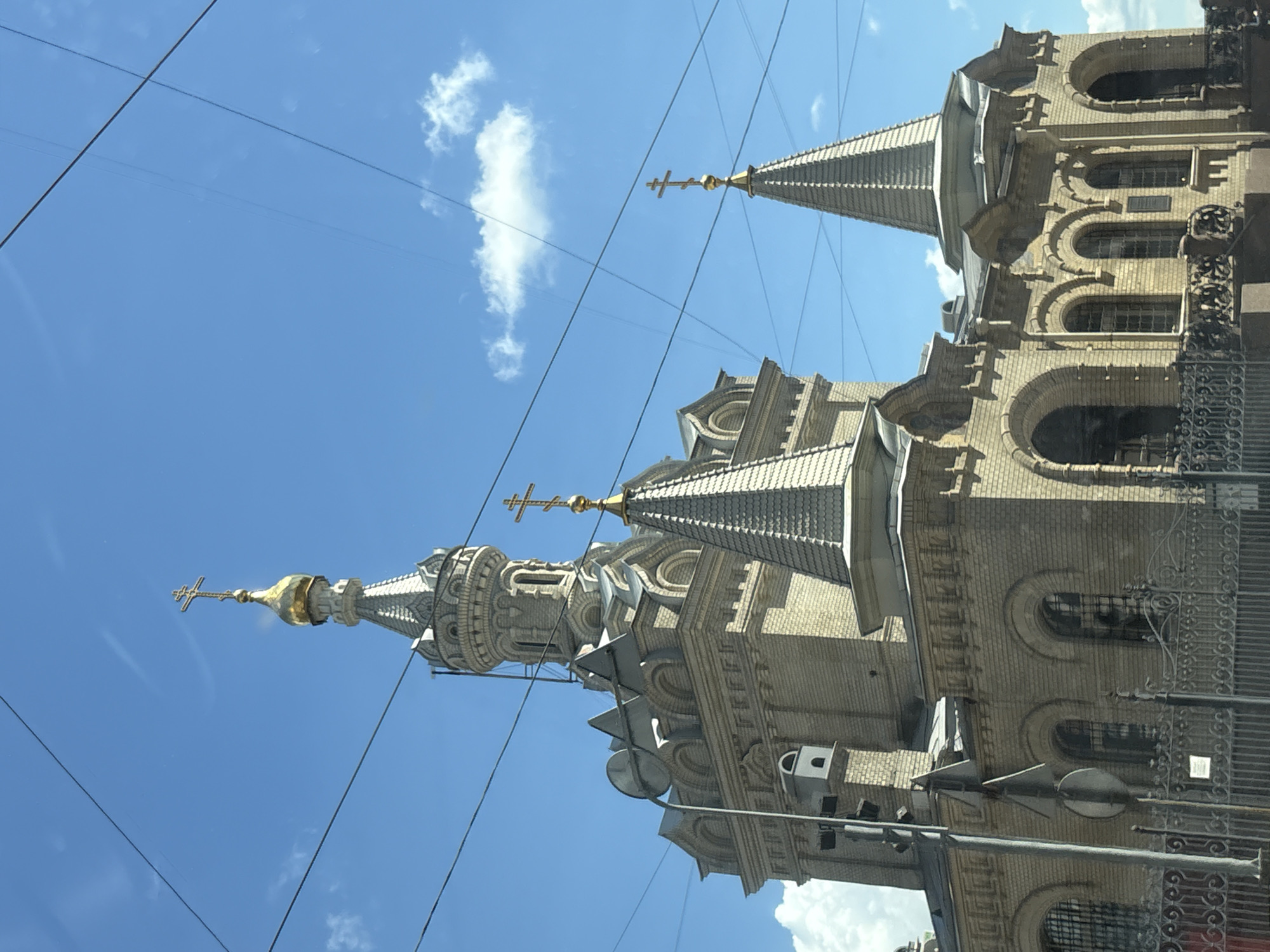 The image shows a light-colored brick building with multiple conical roofs topped with gold crosses, characteristic of Russian Orthodox churches. The main structure is tall and ornate, with arched windows and decorative brickwork. In the foreground, a black metal fence partially obscures the lower part of the building. Overhead, several power lines crisscross the bright blue sky, which has a few scattered white clouds. The architectural style and prominent onion domes suggest this is a religious building located in Saint Petersburg, Russia.