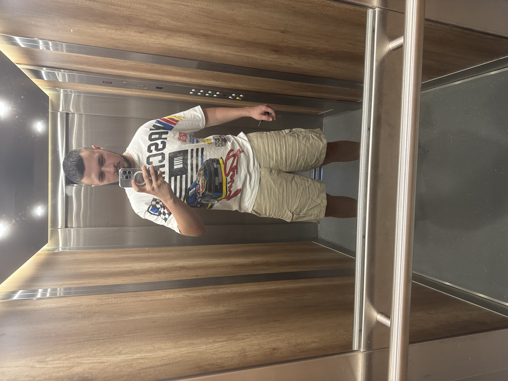 A man stands in an elevator, taking a mirror selfie with his phone. He is wearing a white t-shirt with a graphic print and light brown cargo shorts. The elevator has wooden paneling on the walls and stainless steel railings. The floor is dark gray. The location is not specified in the image, and therefore cannot be determined to be Sofia, Bulgaria.
