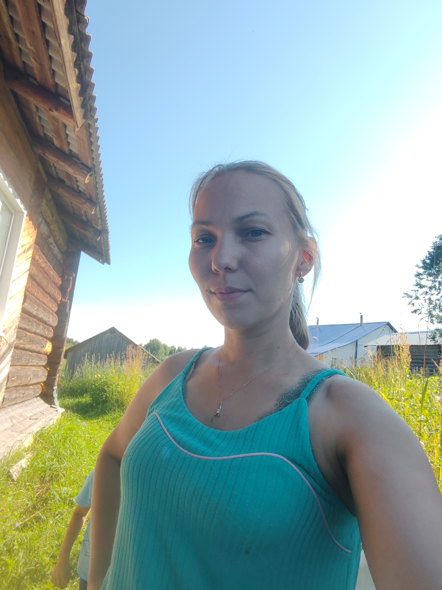 A young woman with blonde hair pulled back in a ponytail takes a selfie outdoors on a sunny day. She is wearing a turquoise tank top with pink trim and a delicate necklace. She is standing in front of a rustic wooden building with a corrugated metal roof. In the background, there are other buildings, tall grass, and trees. The sky is clear and blue. The scene appears to be in a rural or semi-rural area, possibly a village like Чухлэм, Russia, given the architectural style of the buildings and the natural surroundings. A child, partially visible, is walking behind the woman.