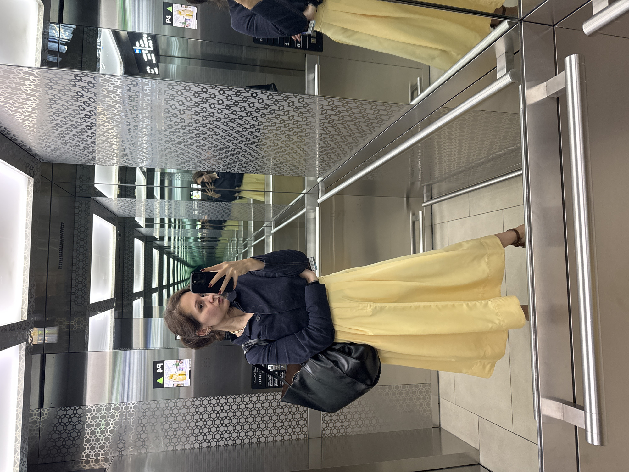 A woman is taking a selfie in an elevator located in Dubai, United Arab Emirates. The elevator has mirrored walls, creating an infinity effect, and is decorated with a geometric pattern. The control panel shows the number 4 with an upward arrow, indicating the fourth floor. The woman is wearing a dark blue jacket and a light yellow dress. She is holding a black smartphone and has a black purse over her shoulder. The elevator interior is brightly lit.