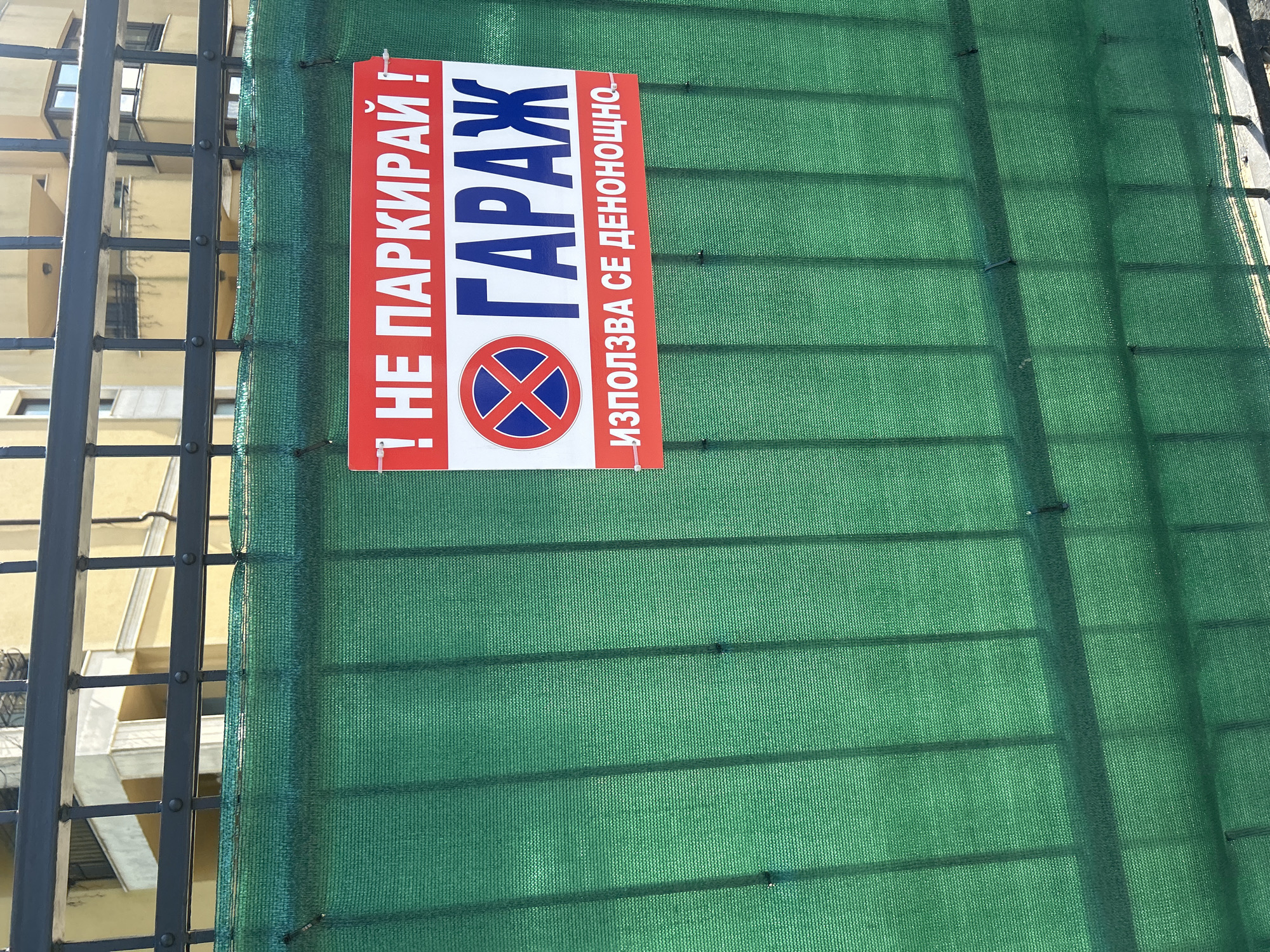 The image shows a sign attached to a green mesh fence. The sign is red and white and contains text and a symbol. The text is in Bulgarian. The top of the sign reads "! НЕ ПАРКИРАЙ!" which translates to "! DO NOT PARK!". Below this, in larger, bold, blue letters, is the word "ГАРАЖ" which translates to "GARAGE". Underneath "ГАРАЖ" is a circular symbol with a red border and a red "X" inside on a blue background, indicating no parking. Below the symbol, smaller text reads "ИЗПОЛЗВА СЕ ДЕНОНОЩНО." which translates to "USED ROUND THE CLOCK.". The sign appears to be located in an urban or semi-urban area, with a building and a metal fence visible in the background. The lighting suggests it is daytime. The overall context implies a restriction on parking in front of a garage that is accessible 24/7.