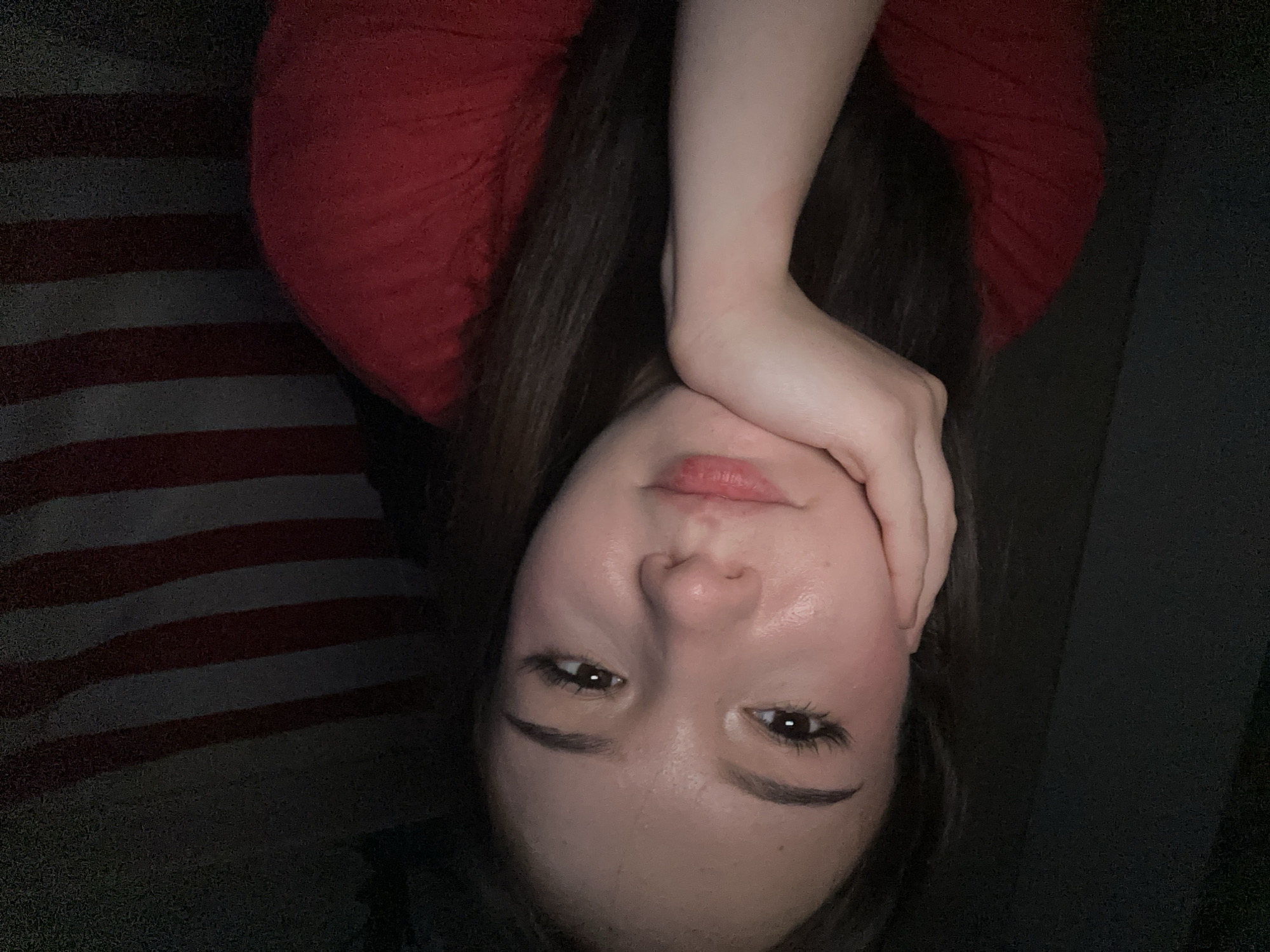 The image shows a close-up of a young woman's face, with her hand resting on her chin. She has long, dark hair and is wearing a red shirt. Her eyes are open and she is looking slightly to the side. The background is dark, with some blurry stripes that appear to be part of a flag, possibly an American flag. The lighting is dim, with some highlights on her face. There is no discernible location in the city of Naberezhnye Chelny, Russia, or any other specific location.