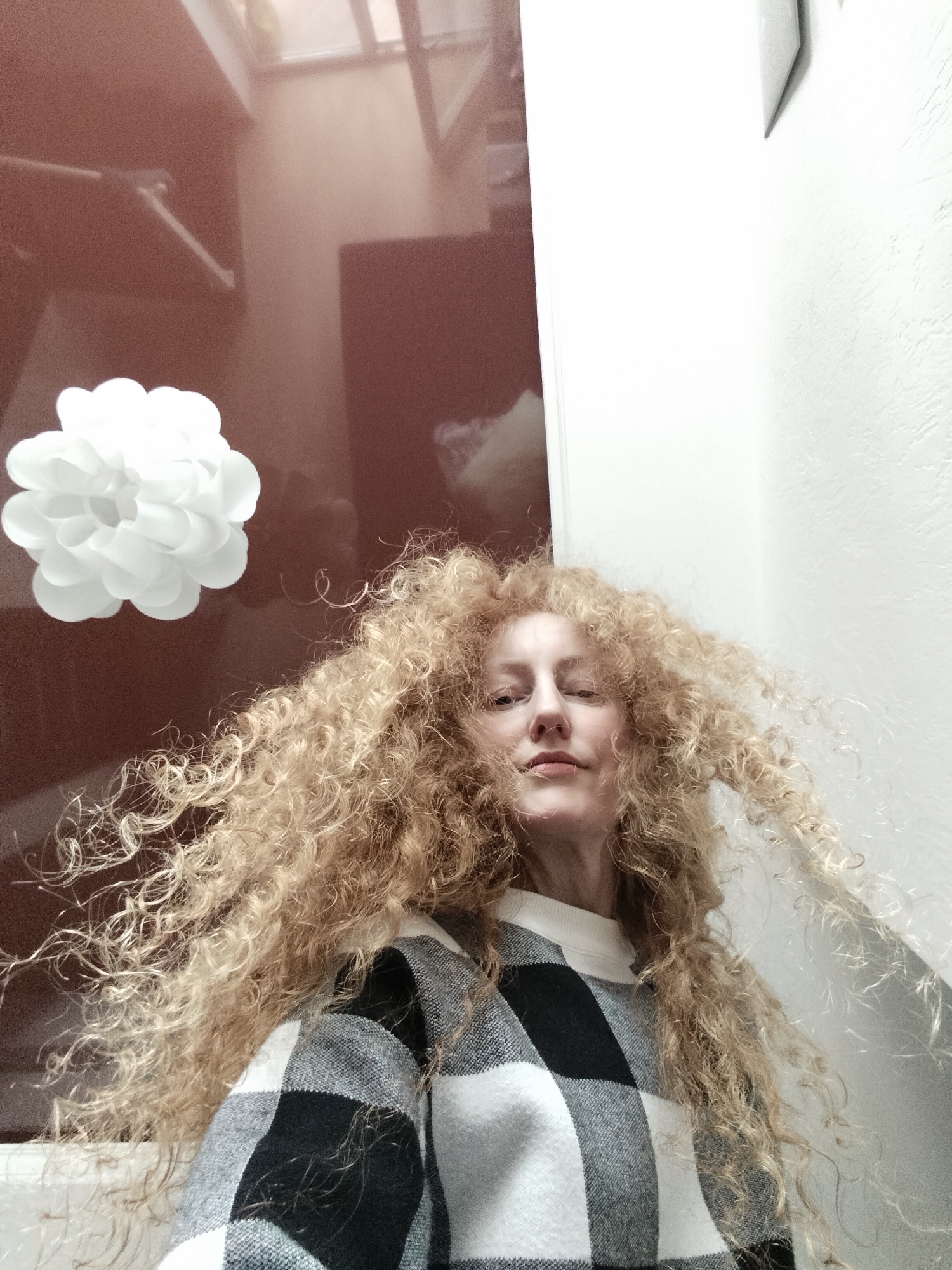 A woman with voluminous, curly, golden blonde hair poses in an indoor setting. She is wearing a black and white checkered sweater over a white turtleneck. A decorative white paper flower is visible on the left side of the frame, affixed to a brown, reflective surface, likely a cabinet. The background also includes a white textured wall on the right, with what appears to be a small picture frame hanging on it. The location or mention of Krykhivtsi, Ukraine, is unknown in the given visual information.