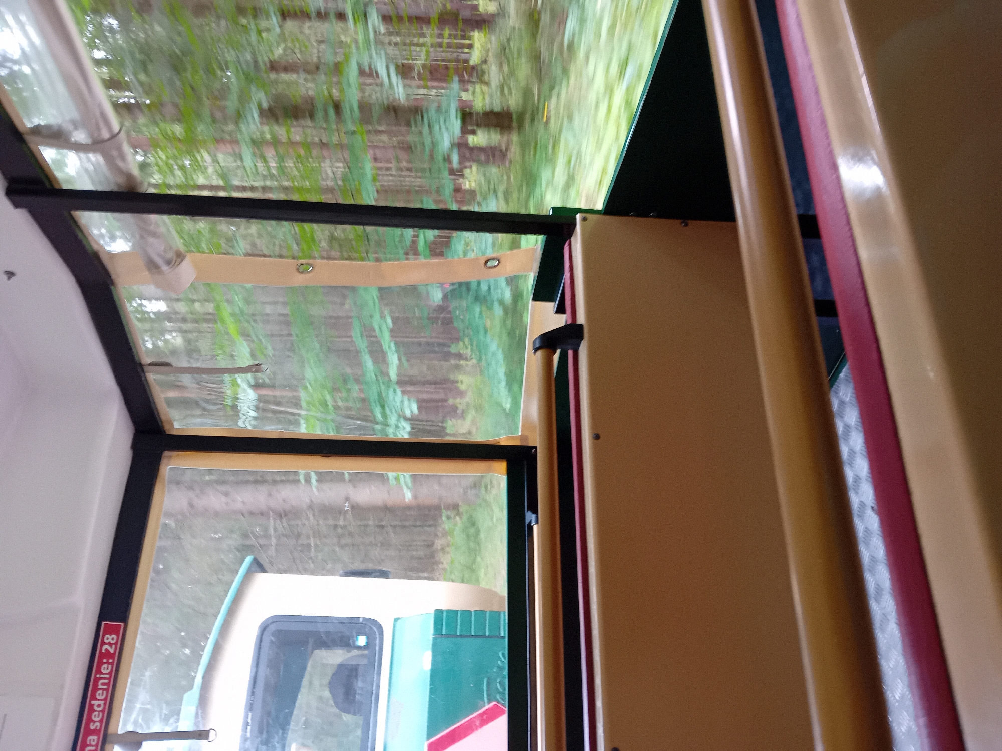 The image captures the interior of a train car traveling through a forest. The view is from the perspective of a passenger, looking out a window. A red and white sign with the text "na sedenie: 28" is visible on the top left of the interior. Inside the train car, there are tan-colored seats with a red trim. The exterior of the train, which appears to be a light beige and green color with a pink and white stripe, is also visible through the window. The forest outside is dense with tall trees and green foliage, suggesting a natural, possibly park-like setting. The motion blur indicates the train is moving. The scene appears to be a leisure or scenic train ride, likely in a forested area near Zuberec, Slovakia, given the context of the question.