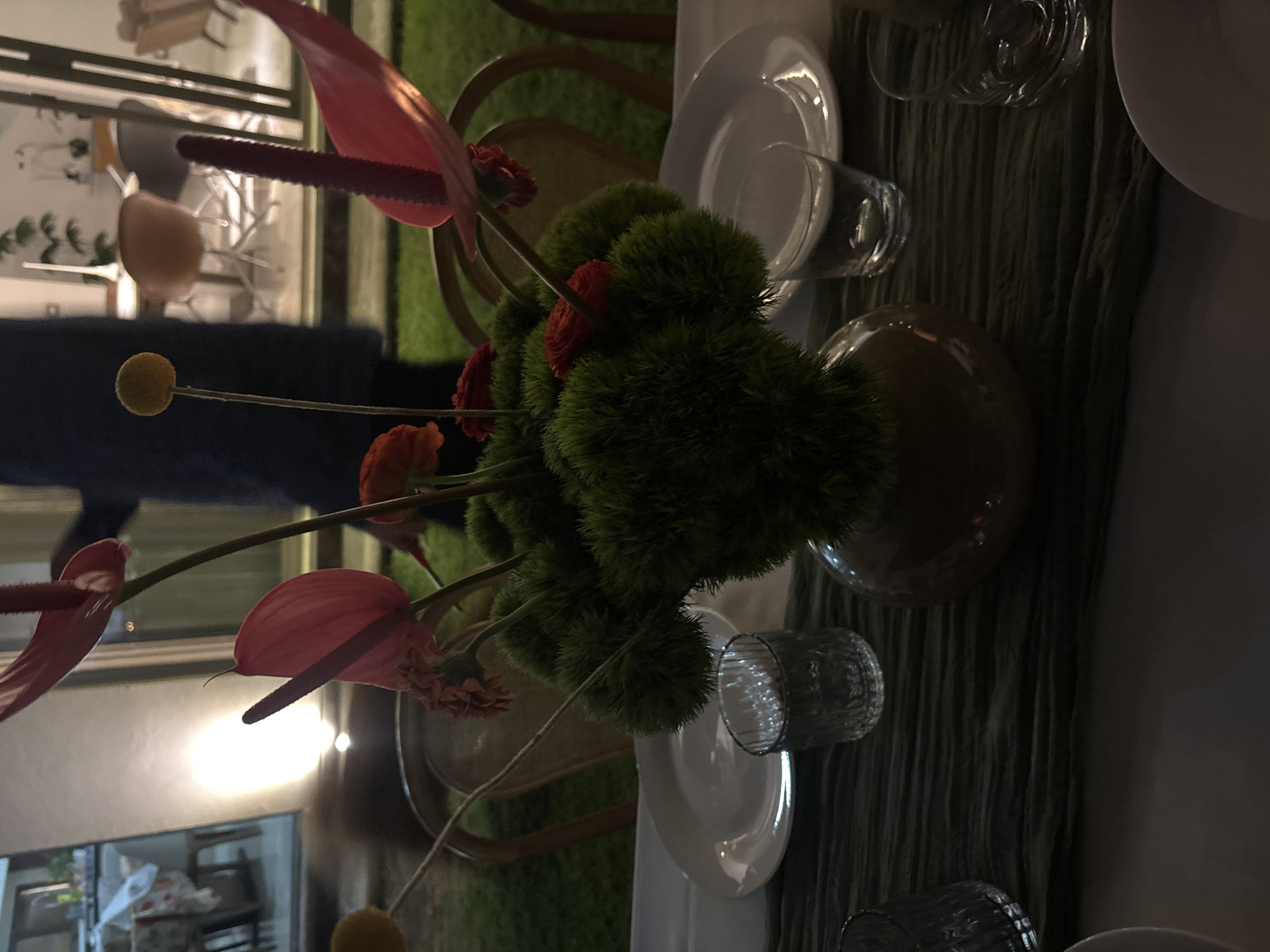 The scene depicts an outdoor evening gathering in the city of Puebla, Mexico, illuminated by artificial light. A table is set with a white tablecloth and a dark olive green table runner. Centrally located on the table is a dark brown, spherical vase holding a floral arrangement. This arrangement features several prominent pink anthuriums with dark spadices, multiple smaller reddish-orange flowers, and yellow spherical craspedia blooms, all nestled within dense, dark green, spiky foliage.
On the table, two white dinner plates are visible, one to the left and one to the right of the centerpiece. Adjacent to these plates are clear, textured drinking glasses. In the immediate background, wooden-framed chairs are positioned around the table on a grassy lawn. Further back, a large glass partition separates the outdoor area from an interior space. Inside, blurred modern chairs and other furniture are visible under artificial lighting. A darkly clad, indistinct individual stands in the mid-background behind the floral arrangement, partially obscured by reflections and the table setup.