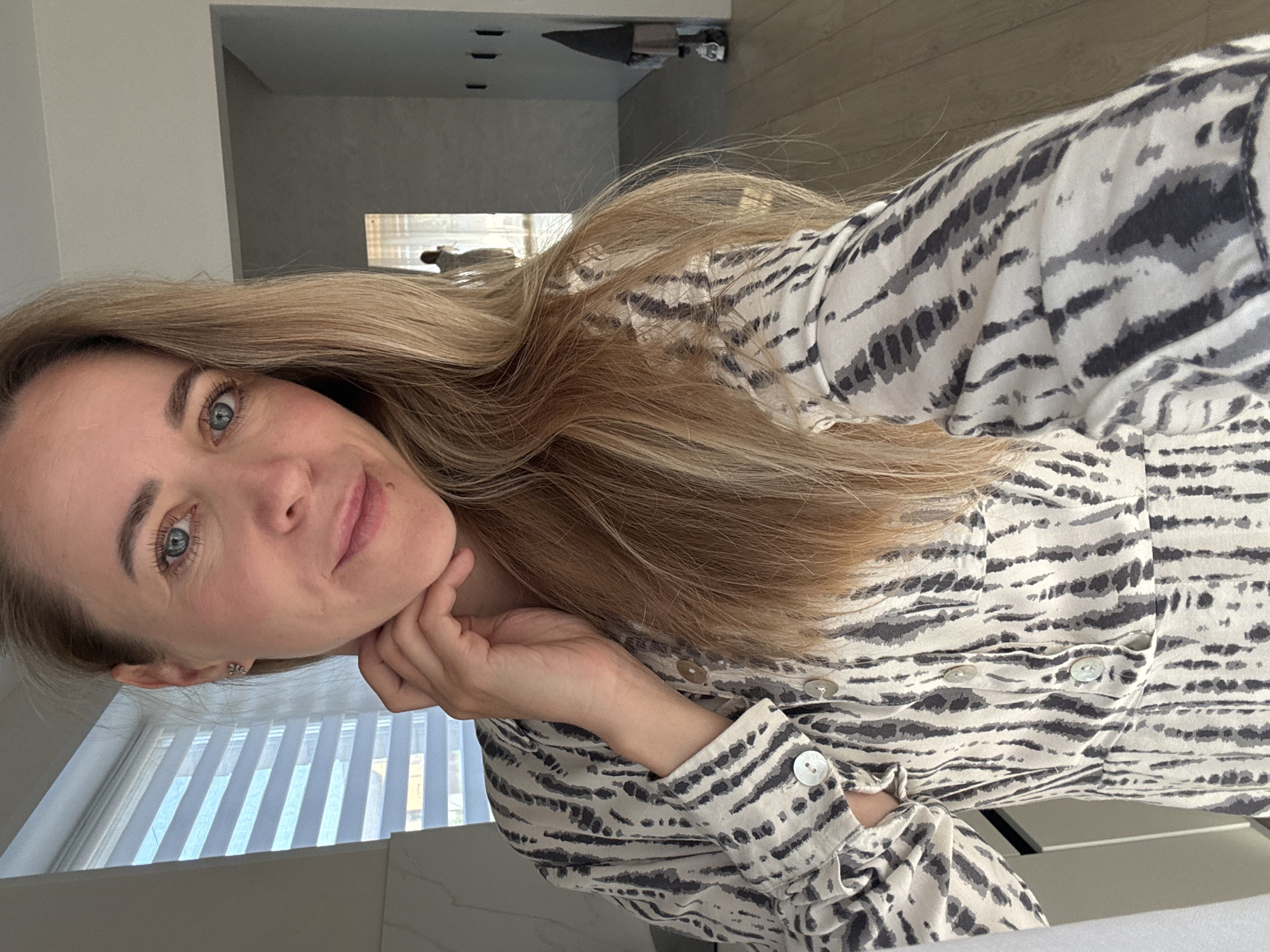 A woman is posing for a photo indoors. She is fair-skinned with light blonde hair and blue eyes. She is wearing a zebra-striped shirt. Her head is tilted, and her left hand is under her chin. The background includes a window with blinds and a glimpse of a room with an open doorway. The location is in Chaikovsky, Russia.