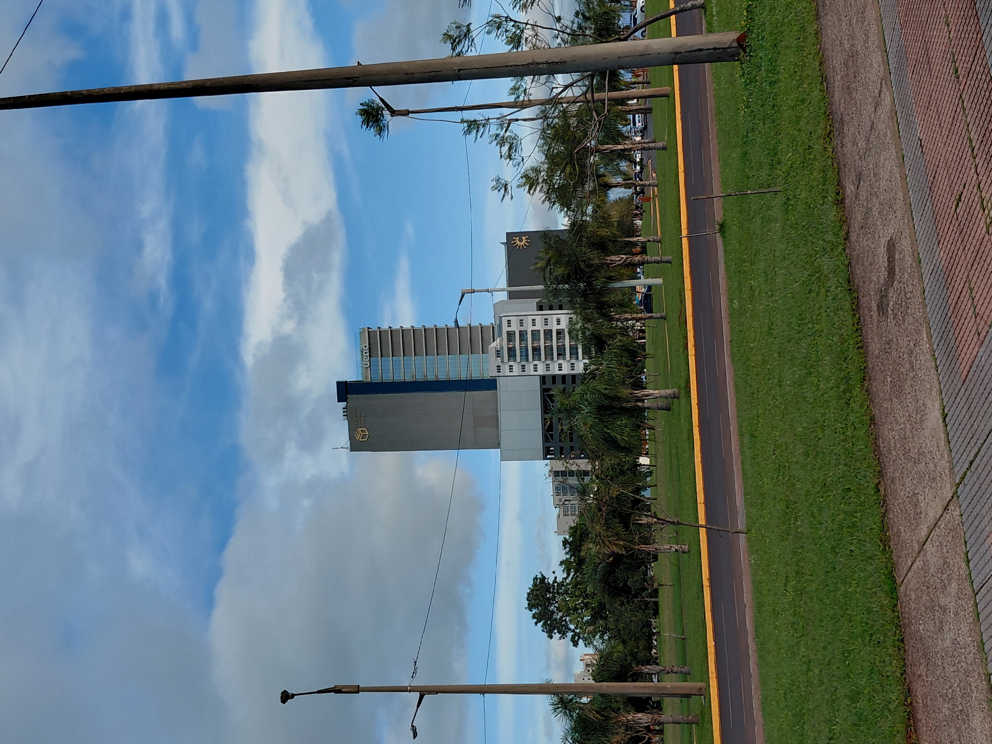 This is a photograph taken from a street-level perspective in Encarnacion, Paraguay. The scene captures a modern architectural ensemble dominated by a tall, multi-story building with a dark grey facade and blue-tinted windows, featuring the logo "VUENO" on its upper section. Adjacent to this structure is another building, lighter in color, with visible balconies and windows. A third, wider building with a dark grey, recessed lower section and a sun-like logo is also visible.
In the foreground, a well-maintained green lawn borders a paved road with a yellow line. Beyond the road, a park-like area is dotted with palm trees and other vegetation. Several lampposts and utility poles line the street, with wires stretching across the scene. In the background, more buildings of varying heights and styles are visible, along with a line of cars on the road. The sky is partly cloudy, with patches of blue visible between the white and grey clouds. The overall impression is one of a developed urban area with contemporary architecture set against a backdrop of green spaces.