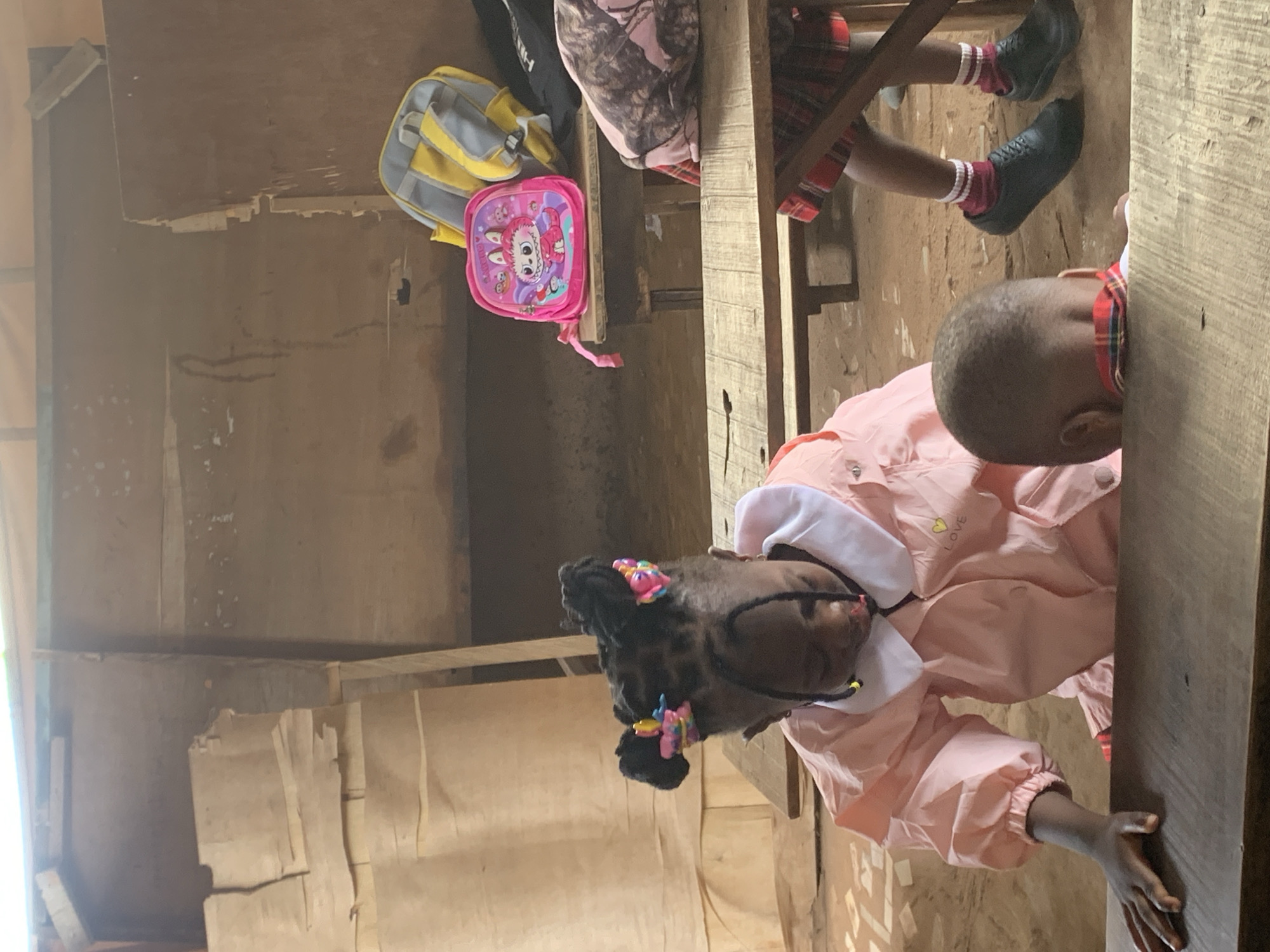 Inside what appears to be a classroom in Ukwu-Abwa, Nigeria, two school-aged children are seated at a wooden table. One child, wearing a light pink uniform, is visible in the foreground, seated with their hand on the table. Another child, in a red and black plaid garment with black shoes and red socks, sits behind them. A yellow and gray backpack and a pink children's backpack are visible on the wooden structure behind the children. The walls appear to be made of wood.