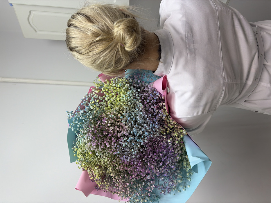 An individual is depicted from the back, standing indoors in a location within the city of Chaikovsky, Russia. The person has blonde hair gathered into a bun at the nape of the neck and wears a light-colored, long-sleeved garment, consistent with a professional uniform or lab coat. They are holding a large, spherical bouquet of small flowers, identified as dyed Gypsophila (baby's breath), exhibiting multiple pastel colors including yellow, green, blue, pink, and purple. The bouquet is wrapped in light blue and pink paper, with a pink ribbon visible on its right side. The background features a plain, light-colored wall. A vertical white pipe or conduit is visible on the left side of the wall, and a white cabinet or door with a recessed panel design is present on the right side.