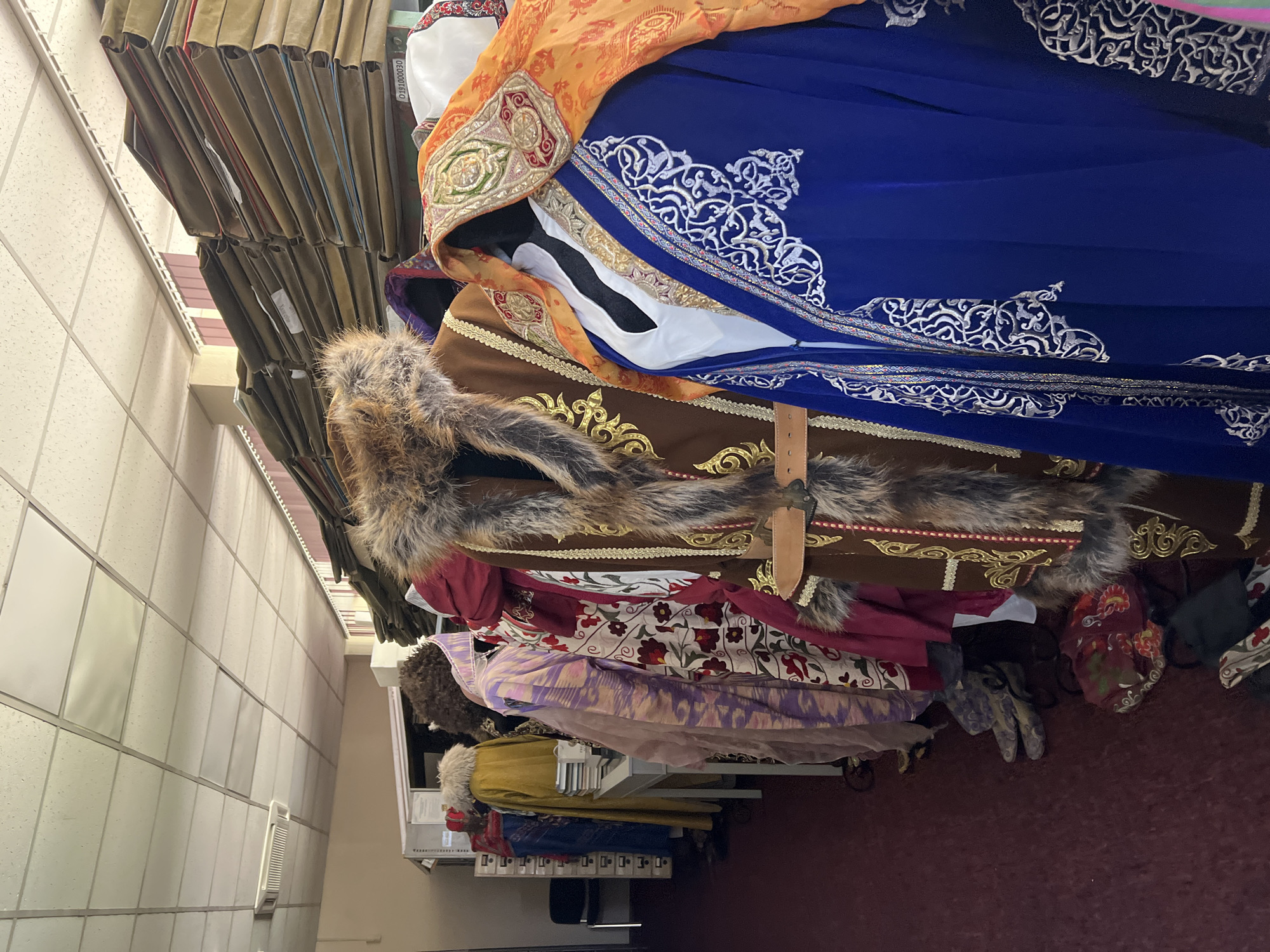 The image shows a row of mannequins dressed in traditional, elaborate clothing, likely on display or in storage. The garments are made of rich fabrics and feature intricate embroidery and fur trim. Behind the mannequins, there are shelves stacked high with what appear to be large documents or files, suggesting a storage or archive area. The environment appears to be indoors, possibly a costume shop, museum storage, or an archive facility. The lighting is fluorescent, typical of an indoor institutional setting. The floor is carpeted in a deep red.