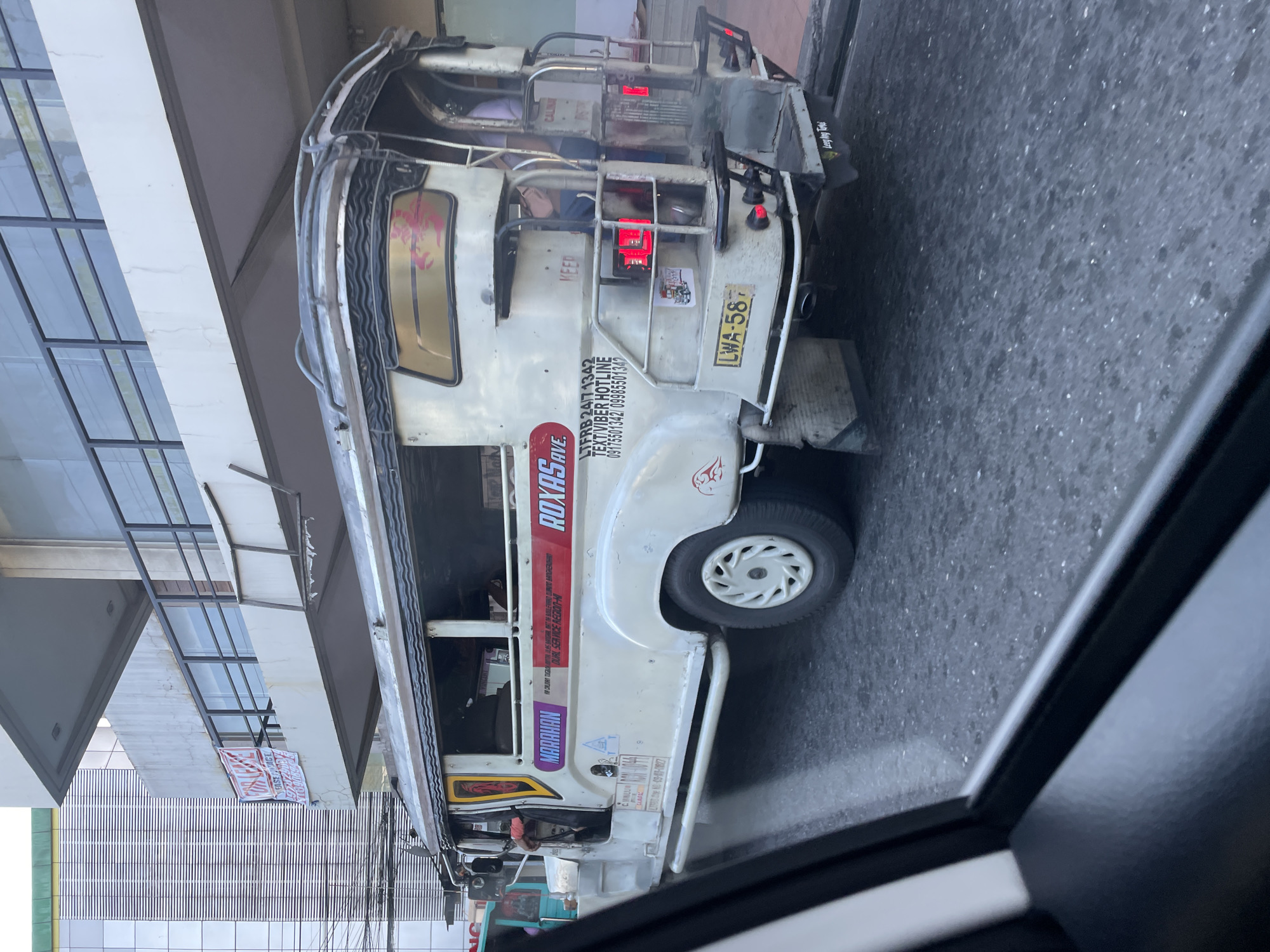 A white public utility jeepney, license plate LWA-58, travels on an asphalt road in Davao City, Philippines. The jeepney displays "MARAHAN" and "ROXAS AVE" as its routes, along with "VIA CALINAN DAVAO-BUKIDNON NATL. RD. VIA DAVAO CITY-BUKIDNON HWY, MARAHAN-ROXAS AVENUE" and "DUAL SERVICE REGION-XI". Contact information for LTFRB (24/7 1342) and a Text/Viber Hotline (09175501342 / 09985501342) is visible on its rear and side. Several passengers are inside, and the driver is visible at the wheel. The jeepney has a roof rack, red taillights, and "Long My Trail" printed on its rear mud flaps. In the background, a multi-story building with a modern glass facade stands to the left. Directly above the jeepney is a concrete building featuring balconies with black railings, from which a red and white "FOR LEASE" sign hangs, partially obscured, with visible text including "FAST...TAGE" and phone numbers "7272101," "09176391191," and "4109106." Overhead electrical wires are visible between the buildings. The image is captured from the interior of another vehicle, with its dark window frame visible in the foreground.