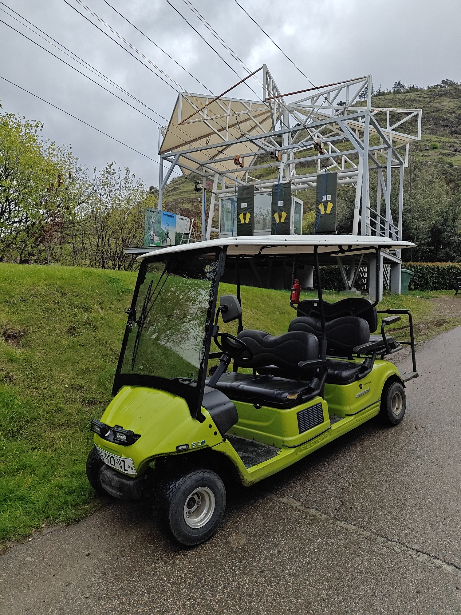 The image captures an outdoor scene at what appears to be a zipline or adventure park in Tbilisi, Georgia, under an overcast sky.
In the foreground, a lime green, multi-seater utility vehicle, resembling a golf cart, is parked on an asphalt path next to a grassy slope. The vehicle has "PILOTCAR" written on its side, "ECO" on the front, and features a visible license plate "N-927-VZ". It has a white roof, a clear windshield with wipers, and four rows of black seats. A red fire extinguisher is mounted inside, near the second row of seats. The asphalt path appears somewhat damp, suggesting recent rain or high humidity.
Behind the vehicle and to the right, a large white metal framework structure dominates the midground. This structure, partially covered with beige canvas awnings, supports multiple parallel cables stretching into the distance, indicative of a zipline launching platform. Several pulleys or rollers are visible on the framework. Below the main platform, there are three dark grey panels, each displaying yellow footprint symbols, suggesting designated standing positions for participants.
Further in the background, a large sign with blue and green graphics is visible through the structure. This sign depicts diagrams of people on a zipline and includes text indicating different lengths: "150 METERS", "500 METERS", and "1200 METER". A partial word "KHUT" can also be seen on the sign. Lush green trees and a rugged, green hillside form the backdrop, suggesting a natural, elevated setting. The overall impression is one of an operational recreational facility ready for visitors. No people are visible, but the setup clearly indicates an activity is about to take place or has recently occurred.