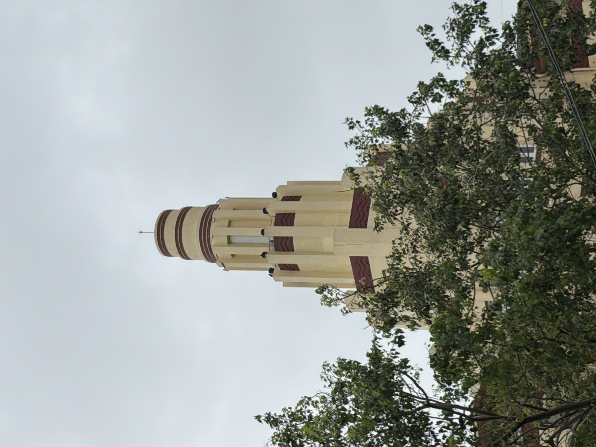 A shot of the Usha Kiran building in Mumbai, India, showcases its distinctive Art Deco architecture against a cloudy sky. The building features a tall, cylindrical tower with horizontal banding in maroon and cream, and decorative maroon zig-zag patterns on its facade. The lower portion of the building, also in cream, is partially obscured by dense green foliage from trees in the foreground.