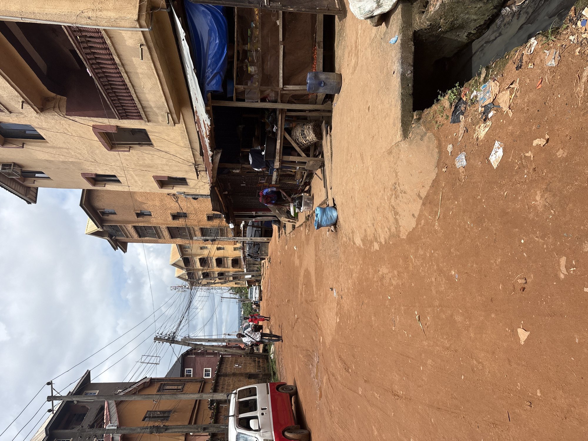 The image depicts a street scene in Nkpor, Nigeria, characterized by a reddish-brown dirt road. Several multi-story buildings, painted in muted earth tones, line the right side of the road. These buildings appear to be residential or commercial, with visible balconies and windows. On the right side of the road, a makeshift stall is set up under a corrugated metal roof with a blue tarp. This stall displays various goods, including produce like tomatoes, and seems to be operating as a small market. A person, partially obscured, is seen working behind the stall.
To the left of the road, a red and white van is parked. Further down the street, a man in a white shirt and dark pants is walking away from the camera. Another person, in a red garment, is walking in the same direction behind him. A few other vehicles are visible in the distance, and utility poles with a complex network of wires are prominent against the cloudy sky. The ground is uneven, with tire tracks visible on the dirt road. There is a drainage ditch on the right side of the road, partially obscured by debris and vegetation, indicating a functional but potentially unmaintained drainage system. The overall impression is one of a busy, lived-in urban environment in a developing area.