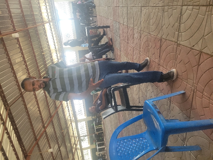 The scene depicts a covered, open-sided gathering area within the city of Ibo, Nigeria. A male individual stands centrally in the foreground, facing right with his head turned left, his right arm extended forward with an open palm as if gesturing. He wears a short-sleeved polo shirt with alternating horizontal stripes of olive green and light gray, blue denim jeans, and dark lace-up sneakers with white soles. His hair is short and dark, and he has a light beard.
To his immediate left, a bright blue plastic chair with a patterned back is positioned. In the mid-ground, a male is seated in a black chair, leaning back, dressed in a dark shirt. Further into the background, multiple individuals are seated at various black tables with corresponding chairs. One person stands near a table on the right where a water bottle is visible. The floor is composed of interlocking reddish-brown and tan pavers arranged in a circular pattern. The overhead structure consists of a corrugated metal roof supported by dark beams. Bright natural light enters the space from the right side, illuminating the background figures and creating some glare. The overall environment suggests an informal meeting, discussion, or waiting area.