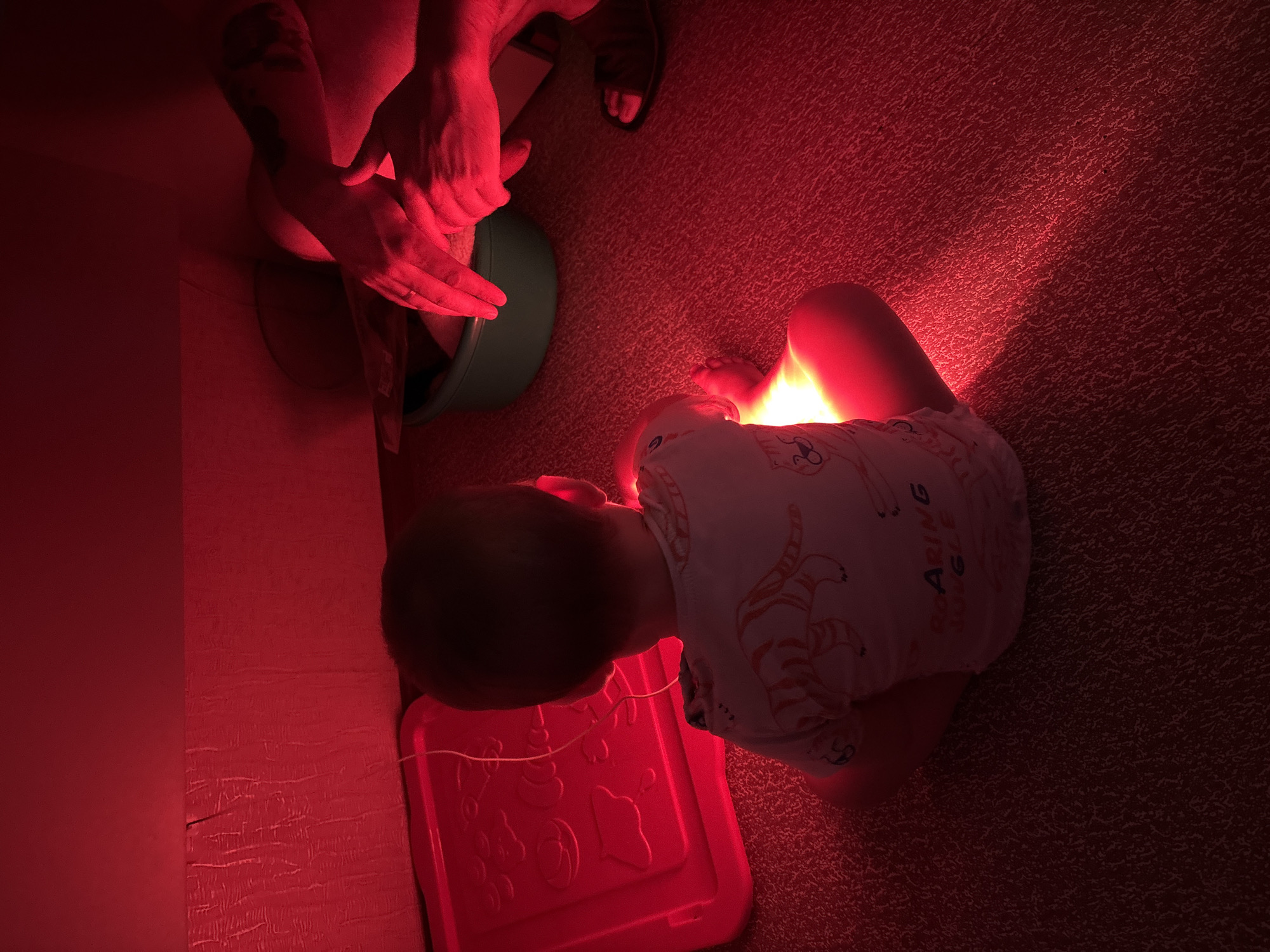 The image depicts a child sitting on a red carpeted floor, facing away from the camera. The child is wearing a white t-shirt with an orange tiger pattern and the words "ROARING JUNGLE" visible. A bright red light emanates from the floor in front of the child, casting a glow on their legs and the surrounding area. To the left of the child, there is a red plastic bin or container with raised patterns. In the background, to the right, there are adult hands, one of which appears to be holding a green basin or bowl. Another adult's foot is visible wearing a sandal. The environment is bathed in red light, suggesting a specific lighting setup. The scene appears to be indoors, possibly a room or a play area. There is no specific indication of the location being in Naberezhnye Chelny, Russia.