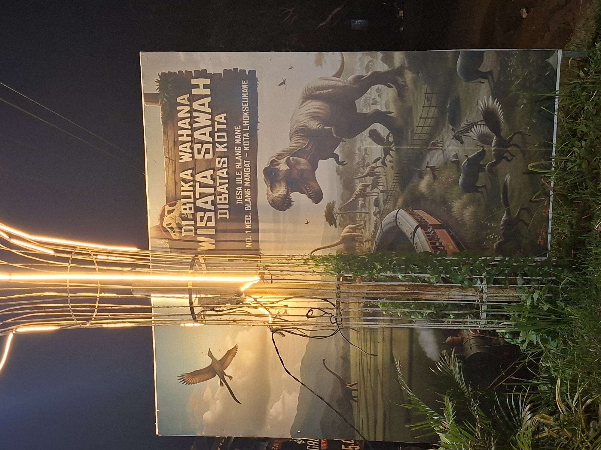 The image shows a sign advertising "Wisata Sawah" (Rice Field Tourism) located on the outskirts of Kota Lhokseumawe, Indonesia. The sign features images of dinosaurs, trains, and birds. The sign is written in Indonesian. It is located at No. 1 Desa Ule Blang Mane, Kec. Blang Mangat. In the foreground, there is an array of bright yellow horizontal lights and greenery.
