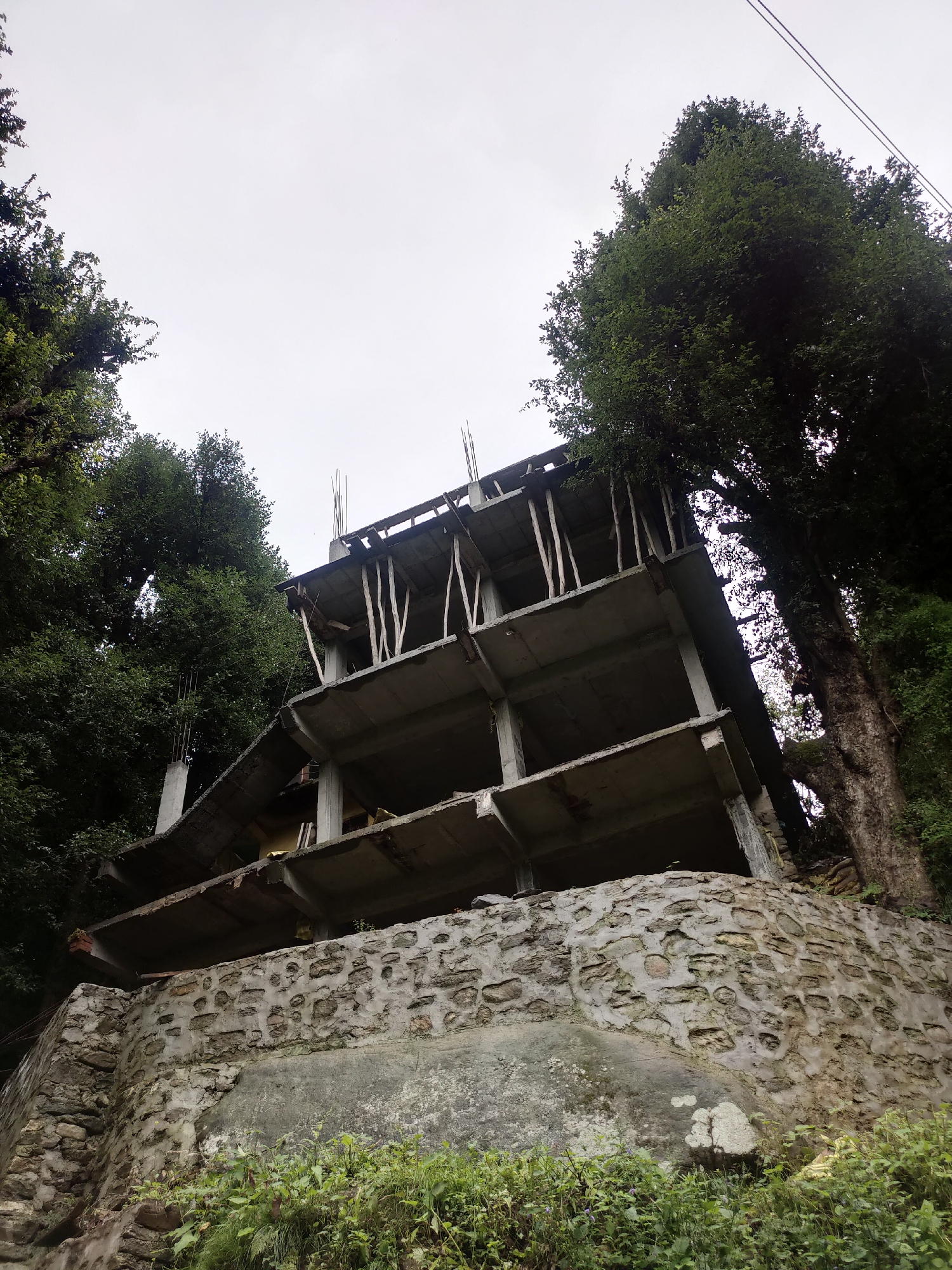 This is a low-angle, eye-level, full shot of an unfinished, multi-story concrete structure located on a hillside in Siyal, India. The structure appears to be under construction, with exposed concrete floors, pillars, and rebar. The building is partially obscured by lush green trees on either side, with a large tree trunk visible on the right. The foreground features a stone retaining wall covered in moss and greenery. The sky is overcast, suggesting a cloudy day. There are no people or animals visible in the image. The overall scene depicts a partially constructed building amidst a natural, wooded environment.