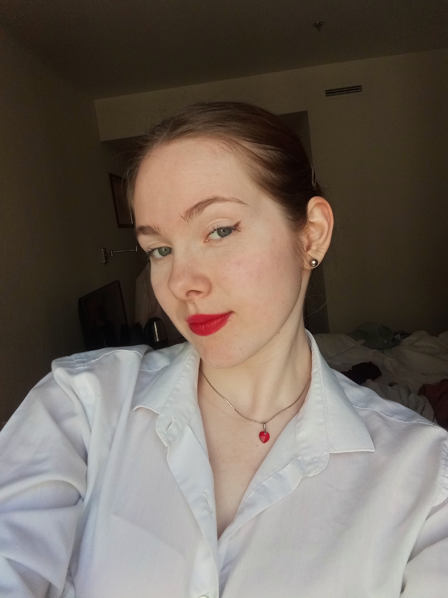 A young woman with fair skin, green eyes, and reddish-brown hair pulled back from her face is posing for a selfie. She is wearing a white button-down shirt, a red heart-shaped pendant on a silver necklace, and a small earring. Her lips are painted with red lipstick, and she has subtle eyeliner.
The background appears to be a dimly lit room, possibly a hotel room. Visible are a wall-mounted air conditioning unit, a framed picture, and a television. There are also rumpled bedclothes. The location in the city of Saint Petersburg, Russia is unconfirmed.
