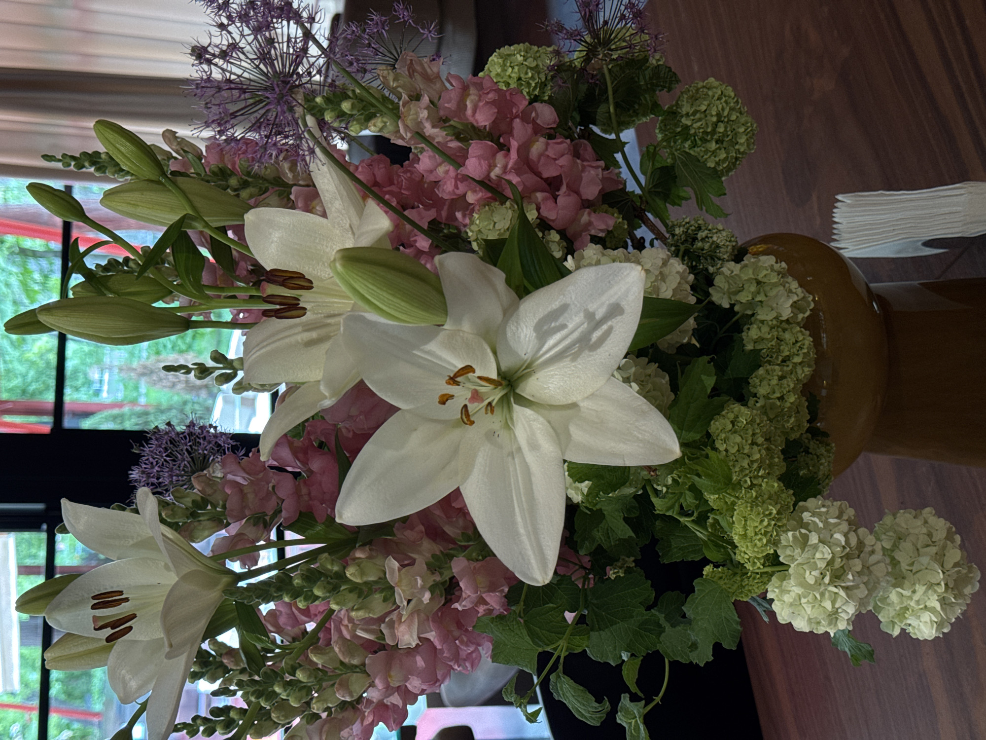 A close-up shot displays a bouquet of flowers in a vase. The bouquet includes white lilies, pink flowers, purple allium flowers, and green hydrangeas. The vase sits on a wooden surface near a window, suggesting an indoor setting. The background shows a blurry view of what appears to be greenery outside. The location of the scene in the city of Yerevan, Armenia, is not verifiable from the image.