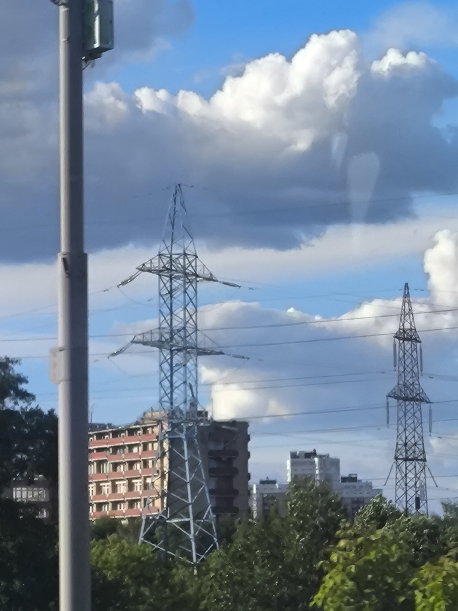 The image depicts an outdoor urban scene under a partly cloudy blue sky. In the foreground, a grey cylindrical pole, possibly a streetlight or utility pole, is visible on the left side. Behind it, a tall, lattice-like metal electricity pylon dominates the center-left of the frame. Further to the right, a second, thinner electricity pylon is visible, also made of lattice metal.
In the mid-ground, several buildings are present. On the left, a multi-story building with a red facade and numerous windows is partially obscured by trees. To the right of this, another, darker building is partially visible. In the distance, a cluster of taller, modern buildings, possibly apartment blocks or offices, can be seen.
The lower portion of the image is filled with lush green trees, suggesting a park or residential area with significant greenery. Power lines are strung between the pylons and stretch across the sky. The overall impression is of a functional urban landscape with elements of both infrastructure and nature. The location is identified as Unknown, Unknown.