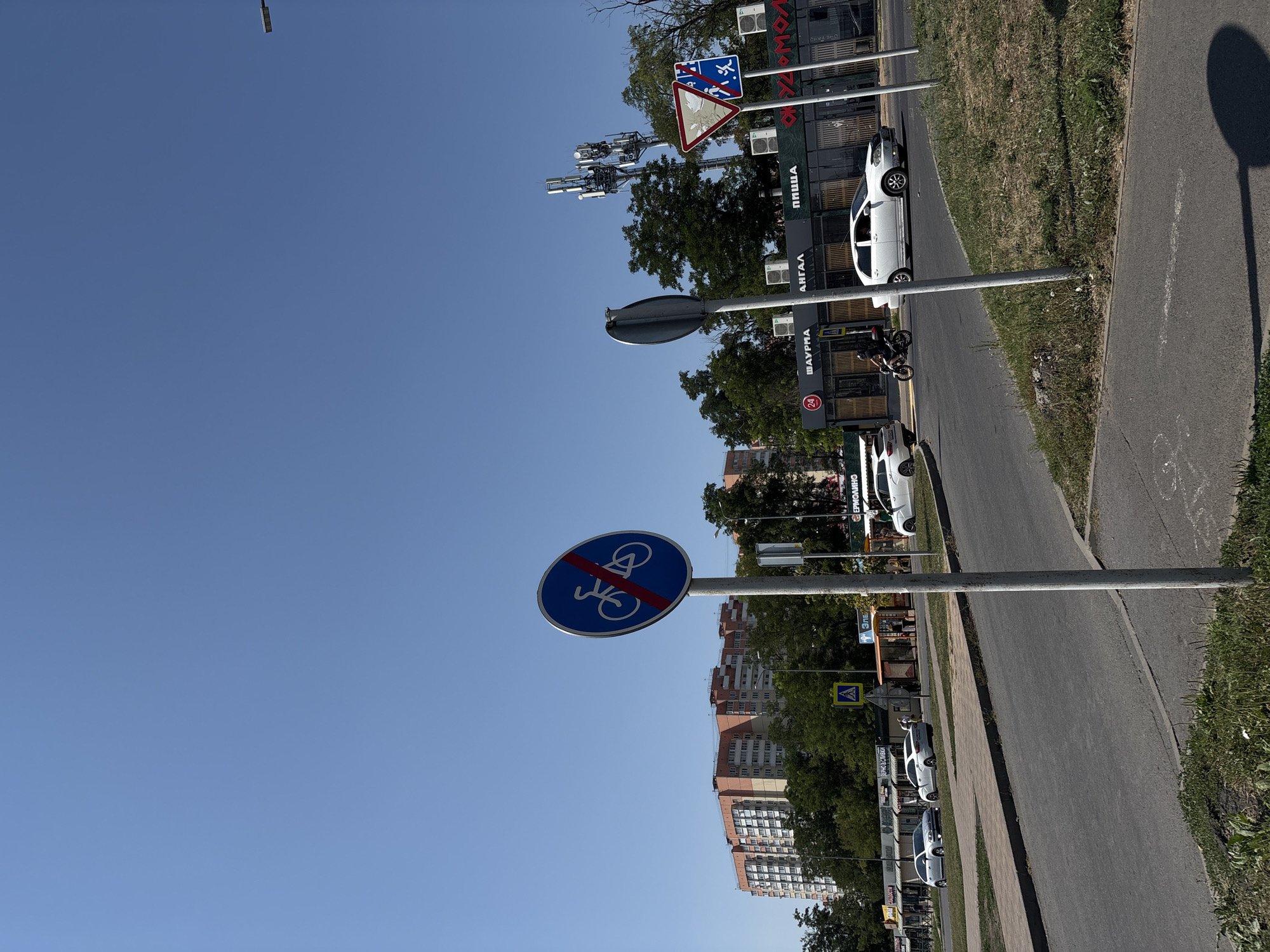 The image captures a street scene in Rostov-on-Don, Russia, under a clear blue sky. In the foreground, a paved road curves to the left, with a grassy verge and a sidewalk. A prominent traffic sign, a blue circle with a red diagonal line through a bicycle symbol, indicates that cycling is prohibited. Further down the road, several cars are visible, including a white sedan and two silver hatchbacks. A motorcycle is also present, with a rider wearing a helmet.
On the right side of the road, a series of shops with awnings line the street. Signs above the shops display text in Russian, including "ШАУРМА" (shawarma) and "ПИЦЦА" (pizza). In the background, tall apartment buildings with light-colored facades are visible, interspersed with trees. Cellular towers stand tall against the sky. Another traffic sign, a red and white triangle with a symbol of a pedestrian crossing, is also present. The overall environment appears to be a suburban or mixed-use urban area.