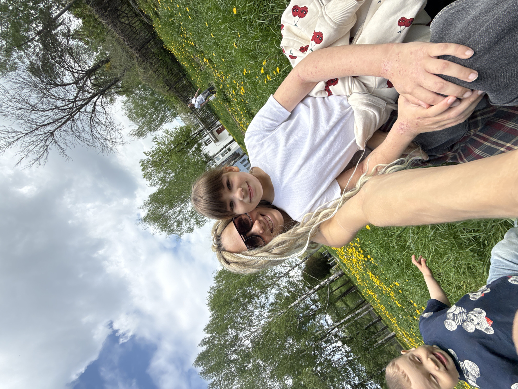A selfie captured outdoors shows a fair-skinned woman with blonde, braided hair and sunglasses, embracing a light-skinned child wearing a white t-shirt. The child is smiling slightly with a head of brown hair. The woman is smiling, and their faces are close. A person with light skin and another child, with short brown hair and a blue shirt patterned with bears, are visible in the periphery of the image. The location is a grassy area with yellow flowers, likely dandelions, under a partly cloudy sky. Trees surround the scene. In the background, a house is partially visible. Given the request context, the location could be in Rusanovo, Russia, but this detail cannot be verified from the image alone. The camera angle is tilted, giving the impression of a self-portrait taken at arm's length.