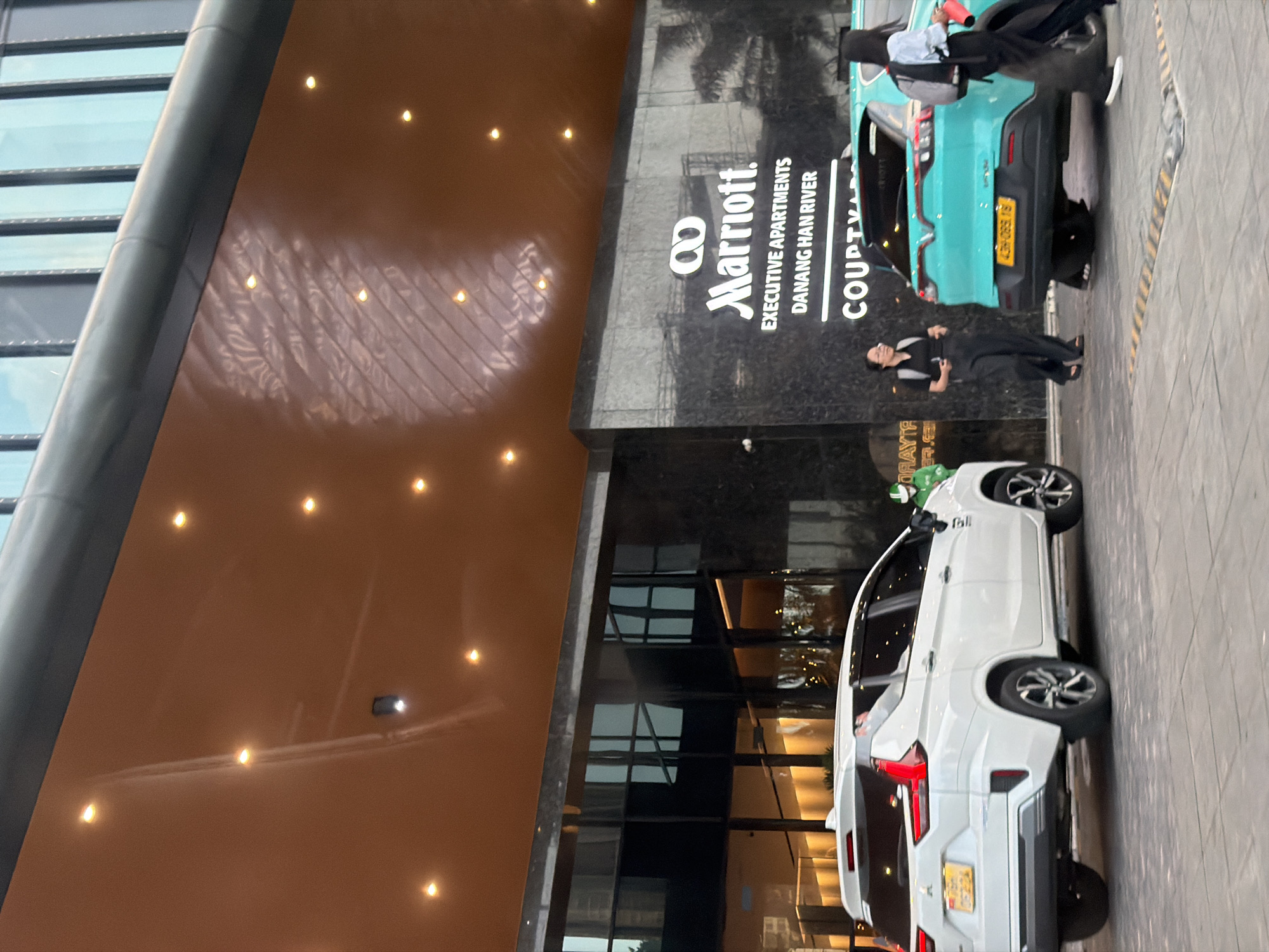 A white Mitsubishi Xpander and a turquoise VinFast VF e34 are parked in front of a modern building with a reflective copper overhang and dark granite facade. The building displays signage for "Marriott Executive Apartments Danang Han River" and "Courtyard by Marriott." Two women are walking near the turquoise car, one in dark pants and a black shirt, the other in dark pants and a light blue shirt. A person in a green delivery uniform is visible to the left of the white car. The scene appears to be a hotel entrance or drop-off area in the city of Đà Nẵng, Vietnam.