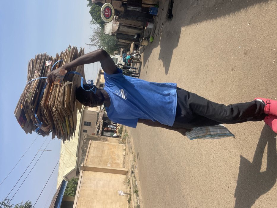 A male individual, appearing to be in his 20s or 30s, stands on a light-colored, possibly asphalt, road in Jos, Nigeria, under clear skies. He is centered in the foreground, facing slightly left from the viewer's perspective. He wears a short-sleeved blue t-shirt with a white logo and text (partially visible: "JD" and "HOMENES"), black trousers, and red clogs or sandals with visible buckles. On his head, he carries a tall, rectangular stack of approximately 20-30 flattened cardboard boxes, secured horizontally with at least two blue plastic straps. His right arm is raised, with his hand lightly resting on the top of the cardboard stack for support. In his left hand, held at his side, he carries a folded, light-colored cloth with thin blue stripes.
The scene is brightly lit by natural sunlight, casting distinct shadows on the road from the individual and objects to his right. The background features a residential street scene. To the left, a light-colored plastered wall (likely a perimeter fence) runs parallel to the road, with parts of buildings and a light-colored roof visible behind it. Overhead, power lines are visible against the sky. To the right, further down the street, various structures are present, including buildings with corrugated metal roofs and walls, indicative of improvised or informal construction. A prominent round "33" Export beer sign, gold and white, is affixed to a pole or building on the right side of the street. Several other individuals are visible in the mid-ground, walking or standing, and a white vehicle (possibly a bus or van) and a motorcycle are also present further down the road.