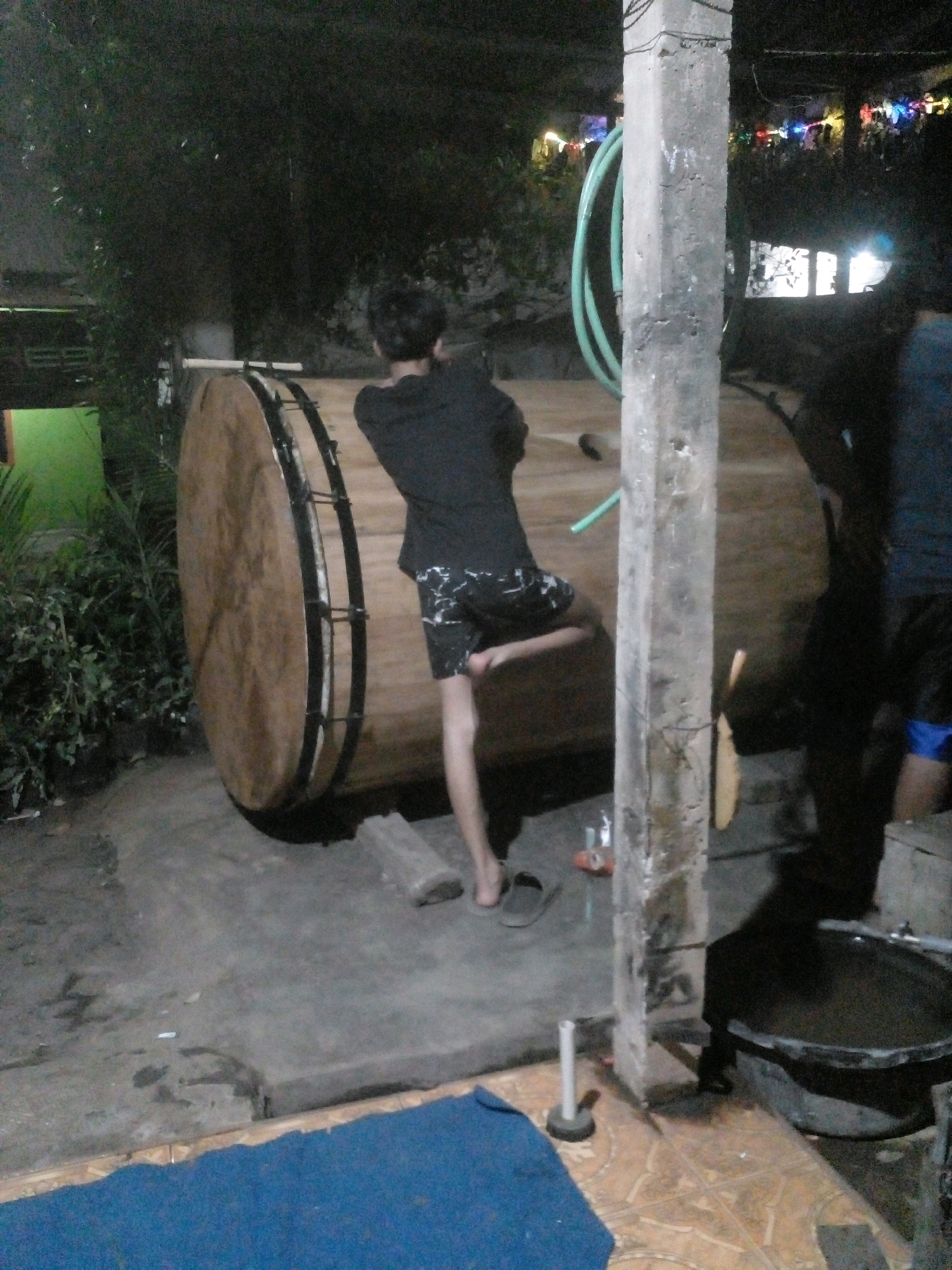 In Pungangan, Indonesia, at night, a person is seen standing next to a large drum. The person is wearing a black shirt and patterned shorts. The drum appears to be made of wood with metal accents. A green hose is coiled nearby. In the background, decorative lights are visible, suggesting a festive occasion. The ground is a mix of concrete and tile. To the right, a large, dark basin is present.