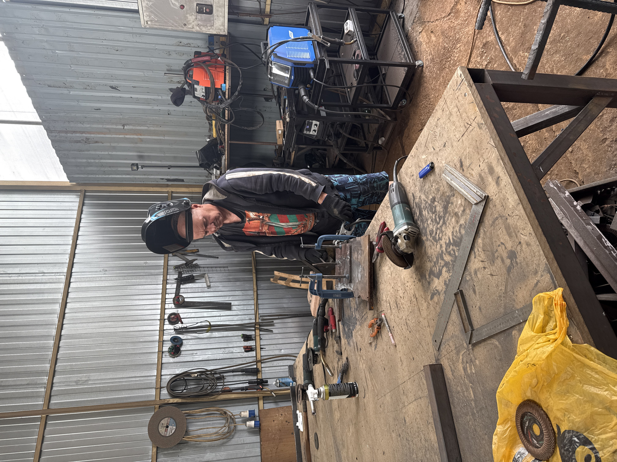 In a cluttered workshop, a man wearing a welding helmet and gloves is standing at a large wooden workbench, appearing to be engaged in welding. The workshop has corrugated metal walls and a visible ceiling with wooden beams. Various tools and equipment are scattered across the workbench and shelves, including welding machines, grinders, clamps, pliers, a torch, and metal pieces. A bright yellow plastic bag with a grinding disc lies on the foreground of the workbench. The floor is dusty and concrete. The lighting suggests it is daytime, with light coming from a window or skylight.