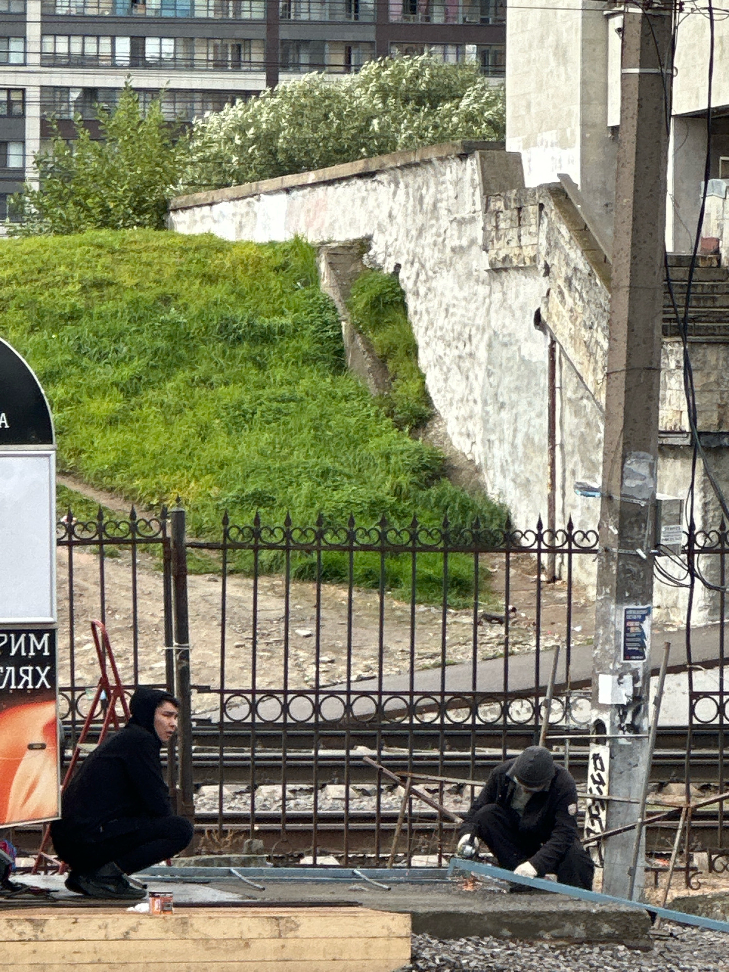 This image depicts an outdoor urban scene in Murino, Russia, likely near a construction or repair site. In the foreground, two individuals are present. One, wearing a black hoodie and black pants, is crouched low, facing left. To their right, a second individual, dressed in a dark beanie, dark jacket, and dark pants, is also crouched, actively engaged in cutting a metal bar with what appears to be an angle grinder, producing sparks. They are wearing white gloves. In front of them are metal bars and debris, suggesting ongoing work.
Behind the individuals, a black wrought-iron fence with decorative circular motifs separates them from a grassy embankment. The embankment is covered in lush green grass and leads up to a weathered, textured white wall. Overgrown greenery is visible behind the wall. In the background, a multi-story building with numerous windows is visible, and further to the right, a concrete utility pole stands with various wires and posters attached to it. A red stepladder is positioned behind the fence near the left side of the frame. Part of a large advertisement or sign is visible on the far left, with Russian text. The overall environment suggests a day with overcast or diffused lighting.