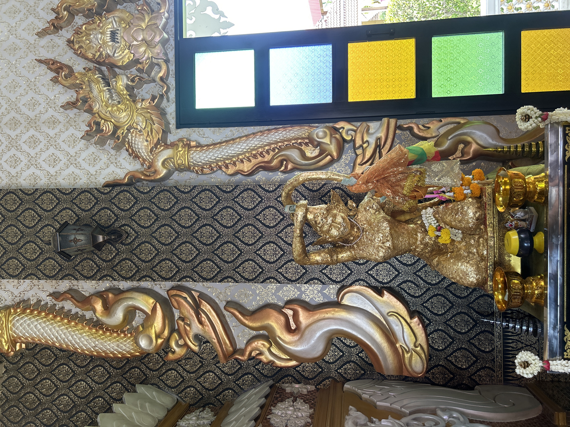 This is a medium shot of a shrine within a building, likely a temple, in Thai Ko Subdistrict, Thailand. The shrine features a golden statue of a deity in a kneeling pose, adorned with a necklace and floral garlands. To the left and right of the statue are ornate, golden, serpentine decorative elements mounted on a dark patterned wallpaper. Above and to the right of the statue, a decorative dragon head is visible. The shrine is situated on a shelf holding various offerings, including golden bowls, a yellow and black alms bowl, and additional floral garlands. Behind the shrine, a window with colored glass panes in green, blue, and yellow is present, suggesting the shrine is located near an exterior wall.