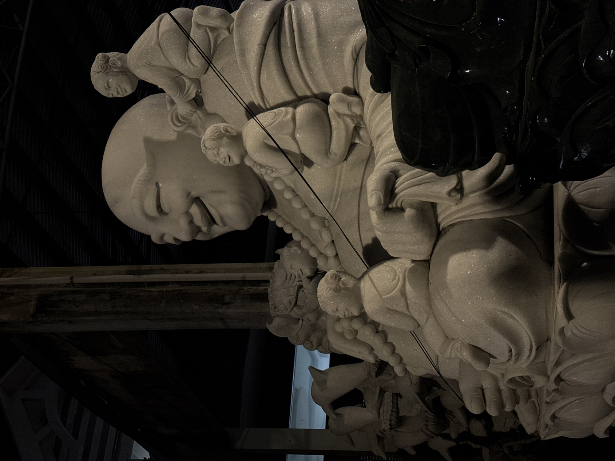 The image shows a close-up of a carved stone statue of a smiling, bald-headed figure with smaller figures clinging to it. The statue is predominantly white or light-colored, possibly marble, and positioned in an indoor setting that appears dim with visible structural elements. There is a dark or black-colored object alongside the white statue. The statue is in Đà Nẵng, Vietnam.