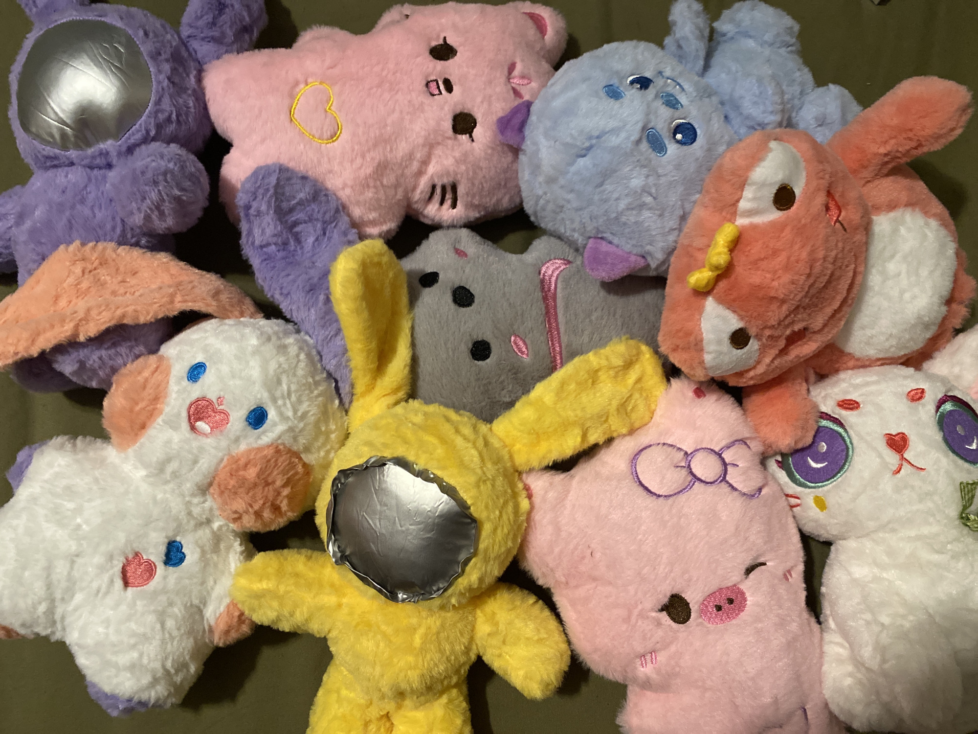 The image shows a close-up, overhead view of a collection of plush toys. There are multiple animal-shaped stuffed animals in various colors, including purple, pink, blue, gray, yellow, orange, and white. Some have decorative features like embroidered hearts, bows, and facial features. The background is a solid, dark green color. The toys are piled together haphazardly. There is no evidence of Khimki, Russia, or any recognizable location. There are no people visible.